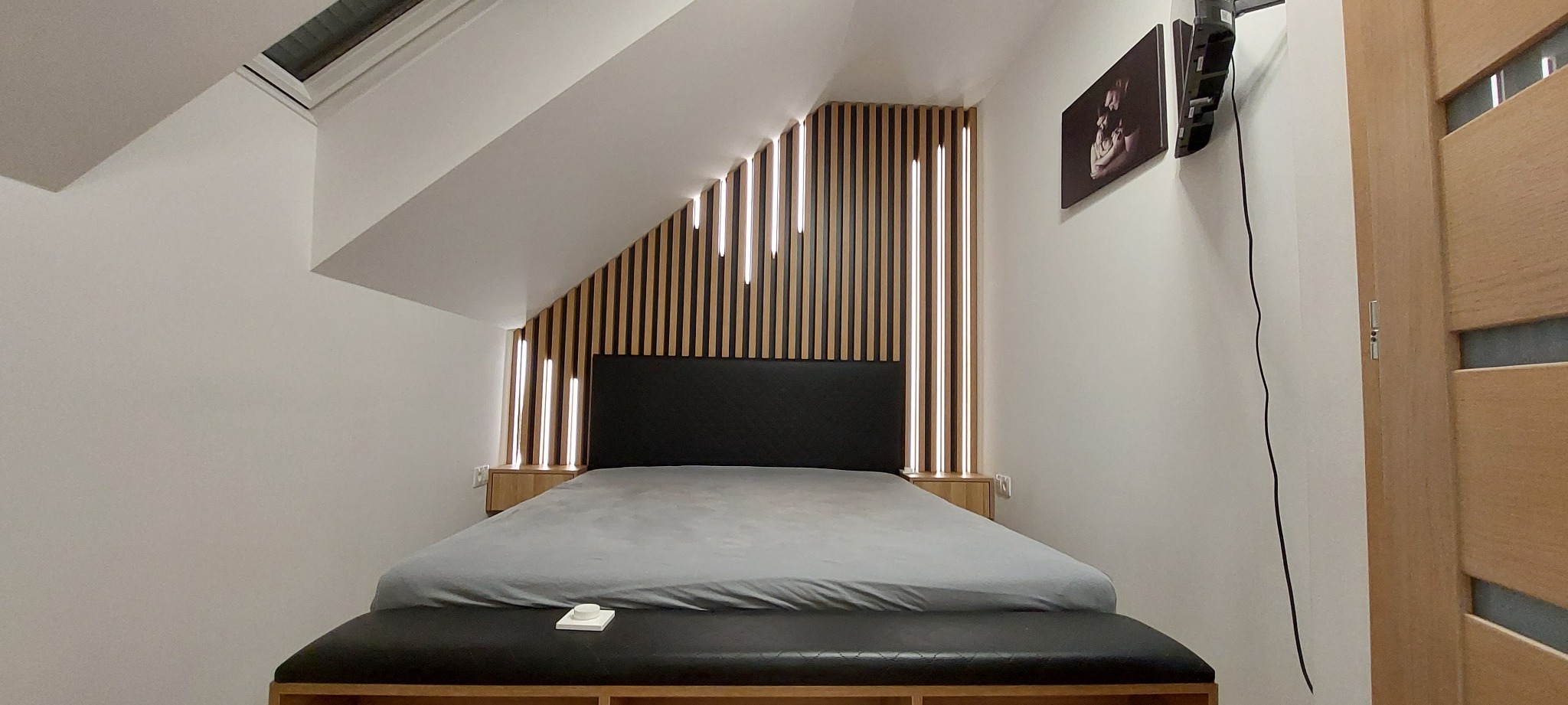 Sypialnia na poddaszu z łóżkiem, drewnianą ścianą z pionowymi listwami podświetlonymi ledami, szafkami nocnymi i czarnym zagłówkiem oraz ławką u podnóża, oświetlona światłem dziennym z okna dachowego.