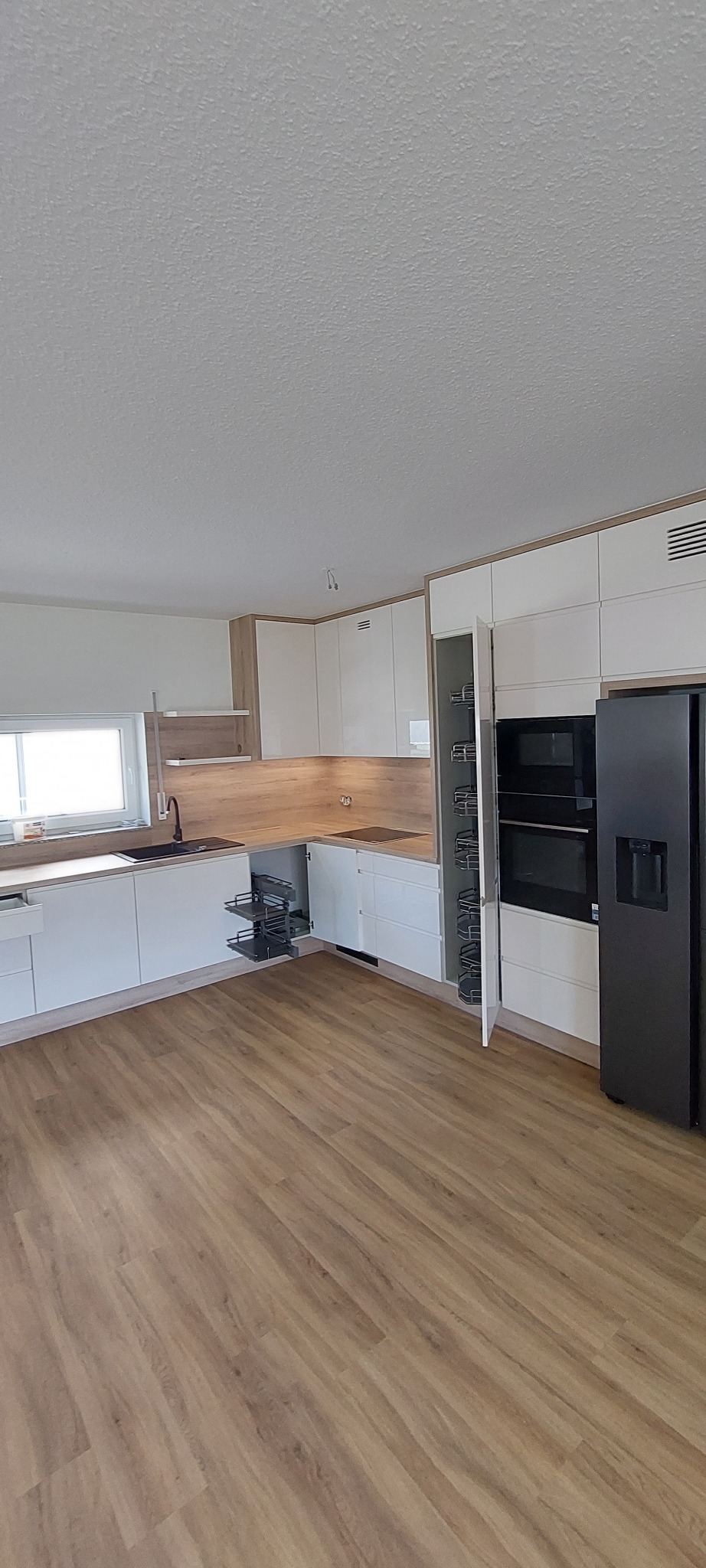 Nowoczesna kuchnia z białymi frontami i drewnianymi akcentami, wyposażona w wysuwane kosze cargo, piekarnik, mikrofalówkę oraz czarną lodówkę, widok na drewnopodobną podłogę.