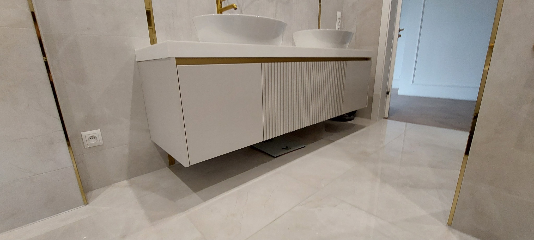 Nowoczesna szafka łazienkowa pod umywalki nablatowe, biała z subtelnymi złotymi akcentami, na tle płytek imitujących marmur i błyszczącej podłogi, z widocznym robotem sprzątającym pod spodem.