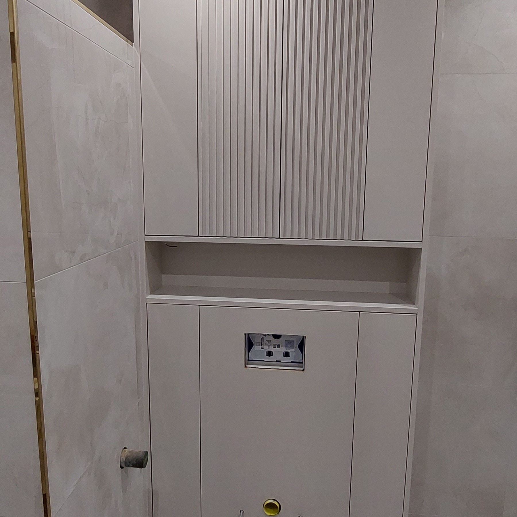 Szara zabudowa meblowa w łazience z szafkami górnymi z pionowymi żłobieniami, półką i dolną szafką z widocznym otworem na stelaż podtynkowy WC.