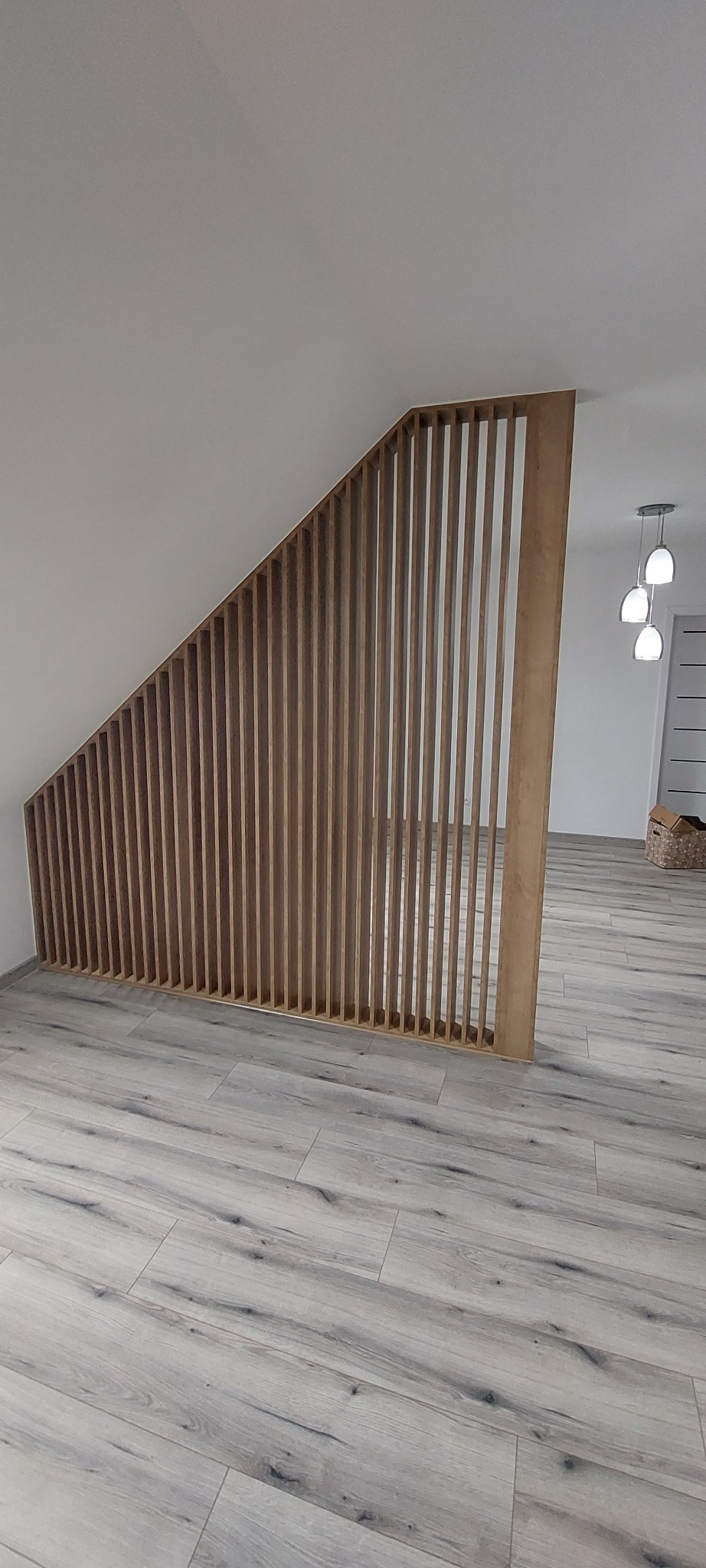 Drewniana konstrukcja z pionowych listew, tworząca ażurową ściankę działową w jasnym wnętrzu z podłogą imitującą drewno, widok na dalszą część pomieszczenia z lampami wiszącymi.