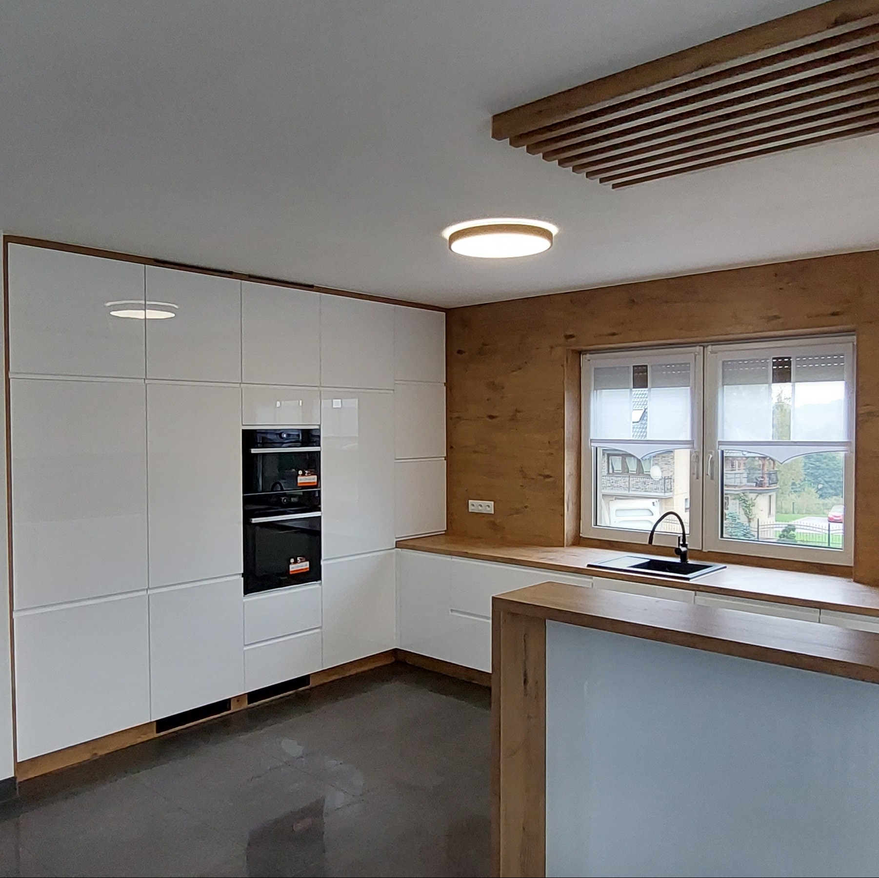 Nowoczesna kuchnia z białymi, lakierowanymi frontami szafek, wbudowanym piekarnikiem, drewnianym blatem i akcentami w kolorze drewna na ścianie i suficie.