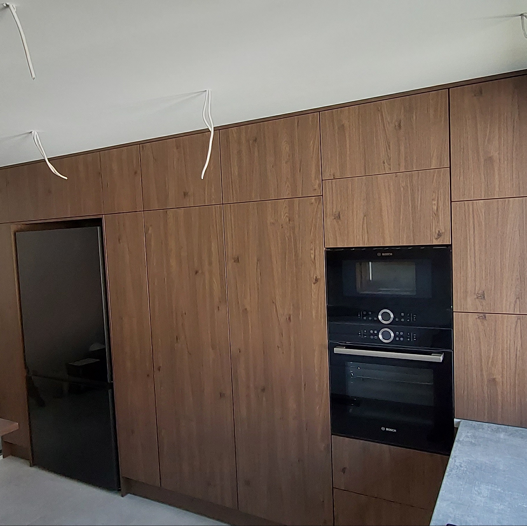 Nowoczesna zabudowa kuchenna z drewnianymi frontami, czarnym sprzętem AGD marki Bosch i wystającymi przewodami elektrycznymi z sufitu.
