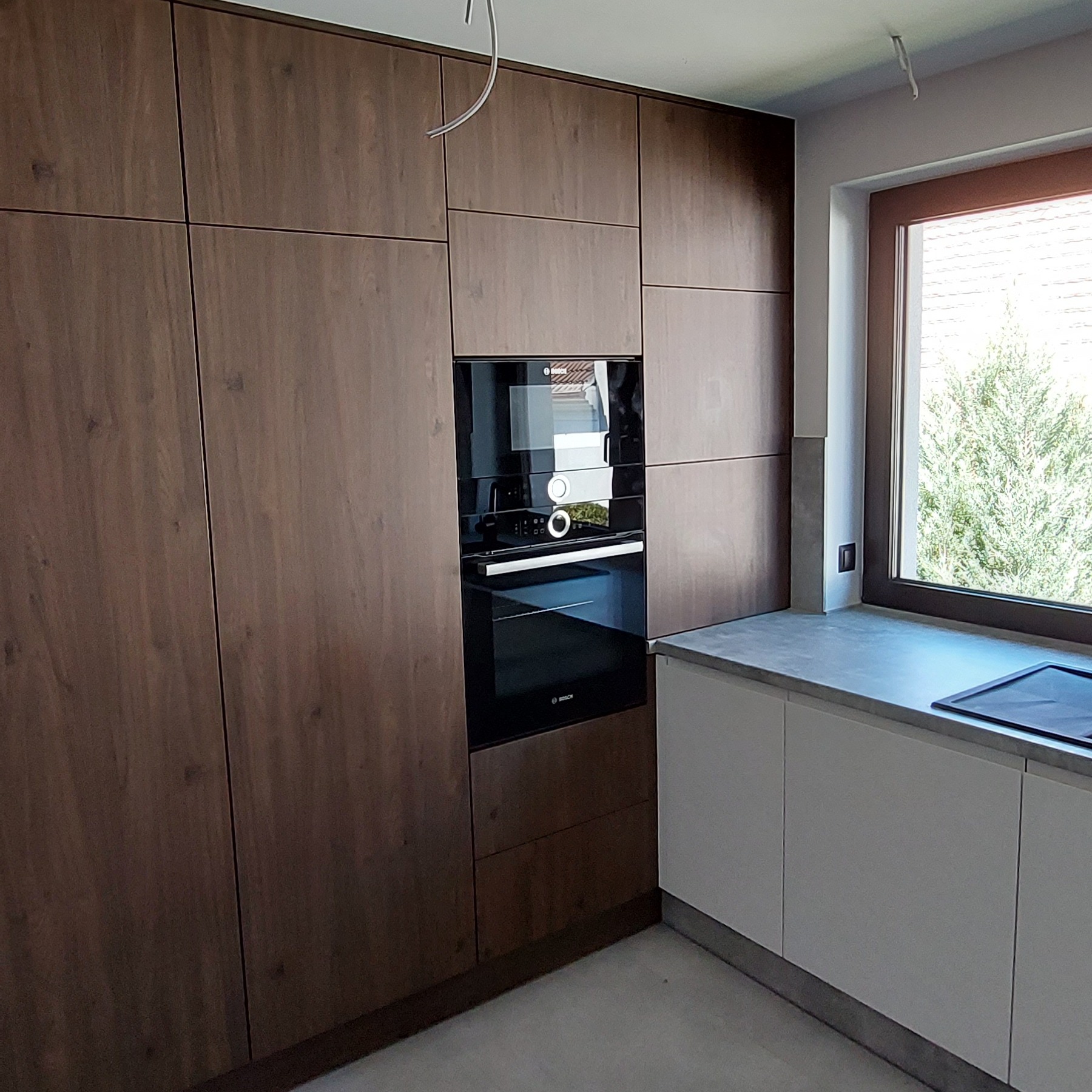 Nowoczesna kuchnia z frontami w odcieniach brązu, zabudowana piekarnikiem i mikrofalówką marki Bosch, jasny blat z oknem w tle, widoczna zieleń drzewa za oknem.