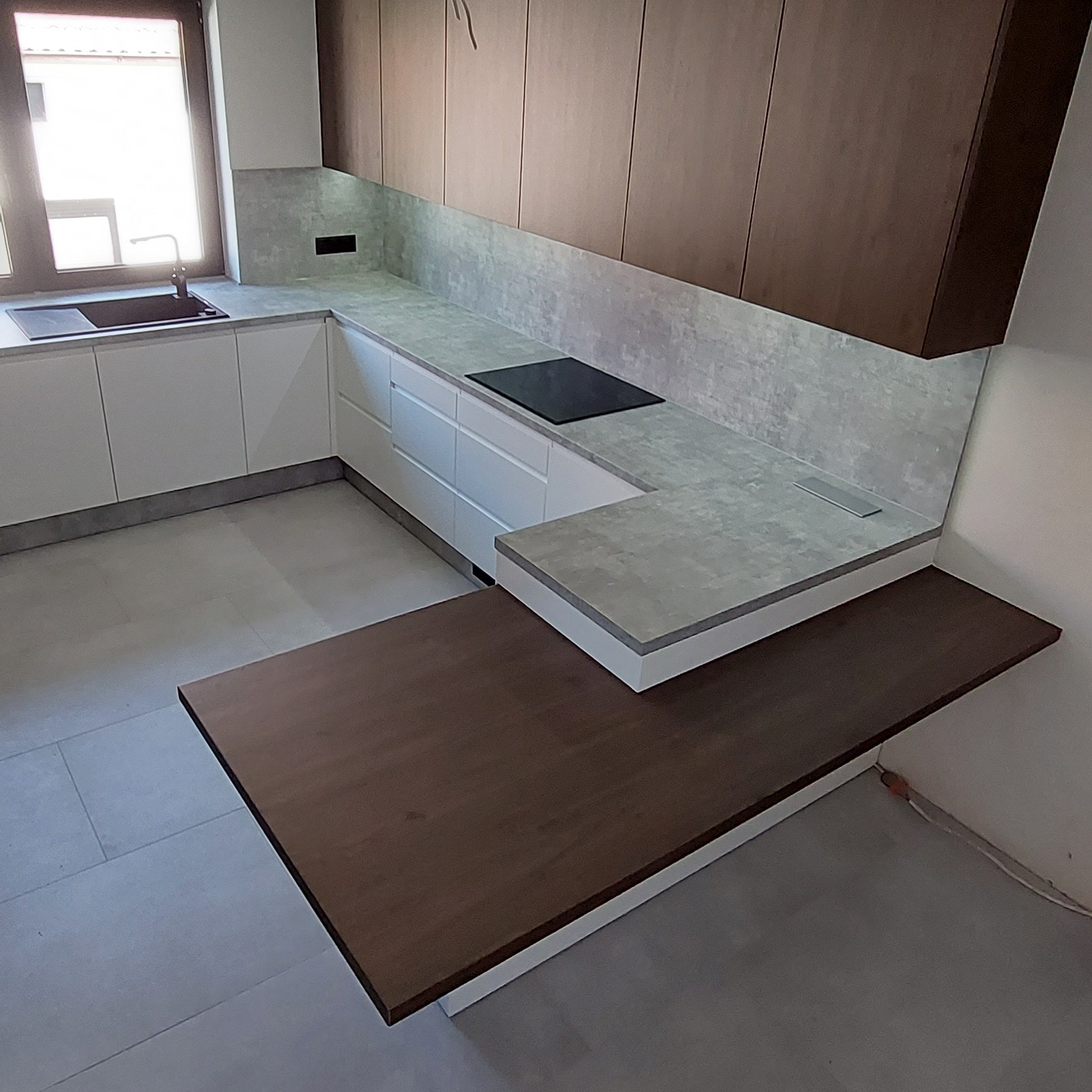 Nowoczesna kuchnia z białymi szafkami, szarym blatem, płytą indukcyjną i drewnianym półwyspem, widok z boku na fragment blatu i półwyspu.