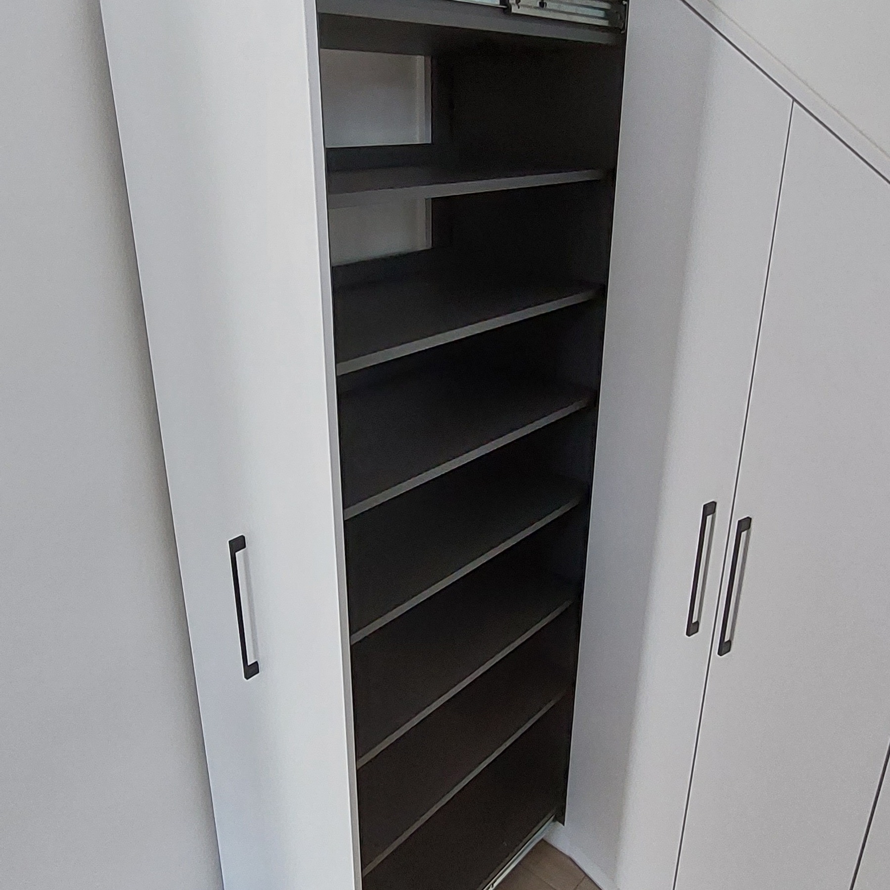 Otwarta szafa wnękowa z szarymi półkami i białymi drzwiami z minimalistycznymi uchwytami, widoczne prowadnice szuflady.