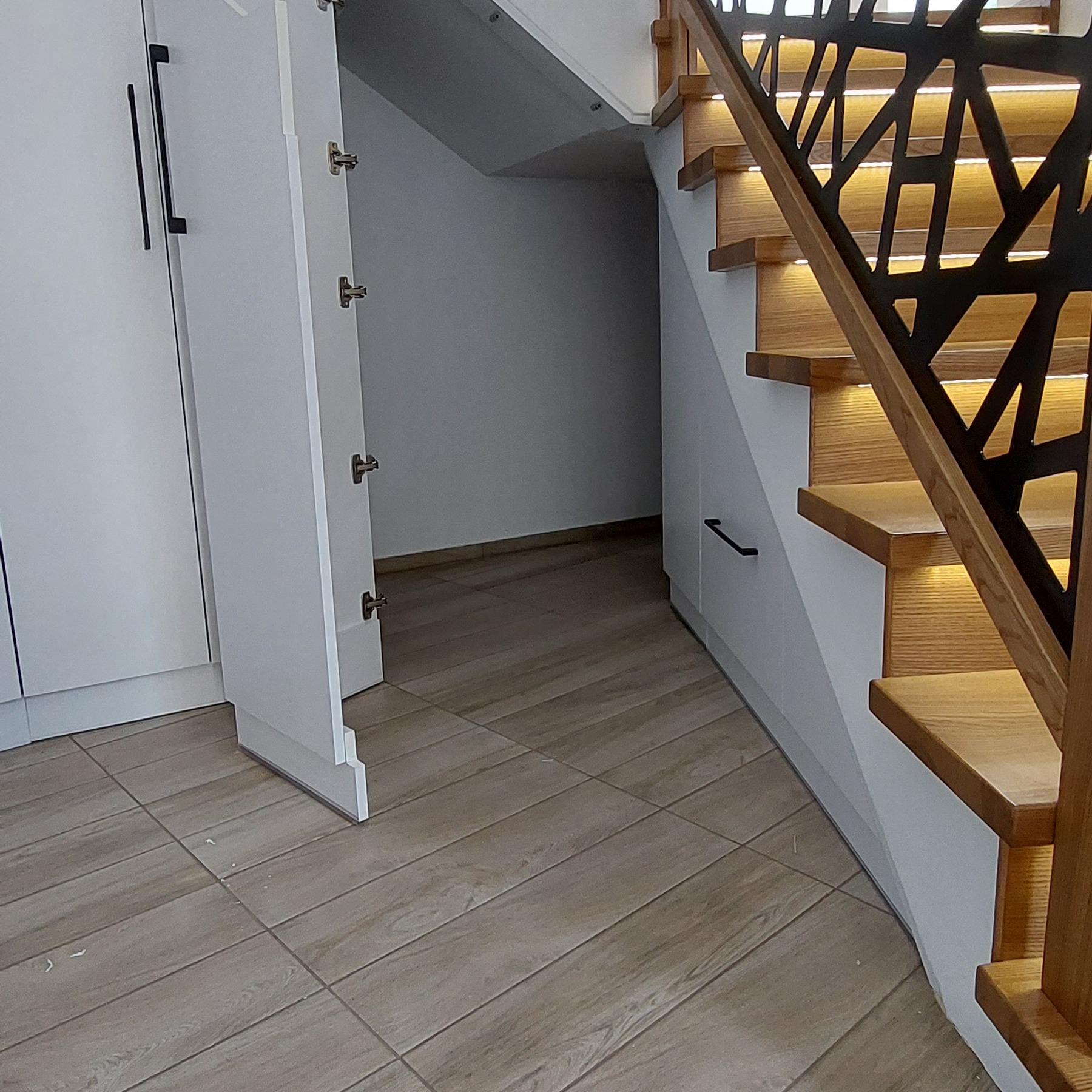 Biała szafa z otwartymi drzwiami obok drewnianych schodów z podświetleniem LED i czarną, geometryczną balustradą.