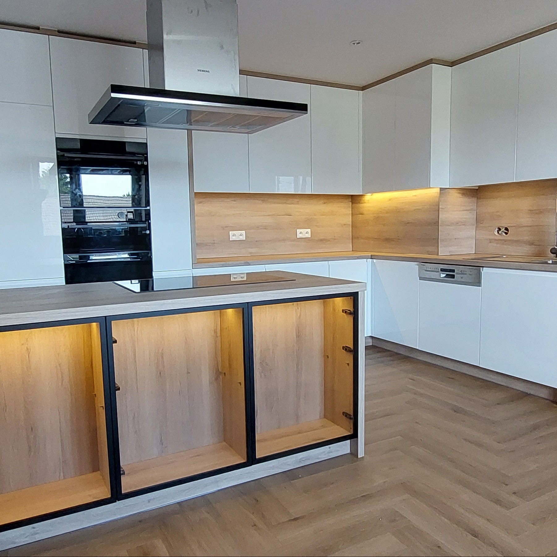 Nowoczesna kuchnia z białymi, lakierowanymi frontami, drewnianym panelem nad blatem i wyspą z oświetleniem LED wewnątrz szafek.