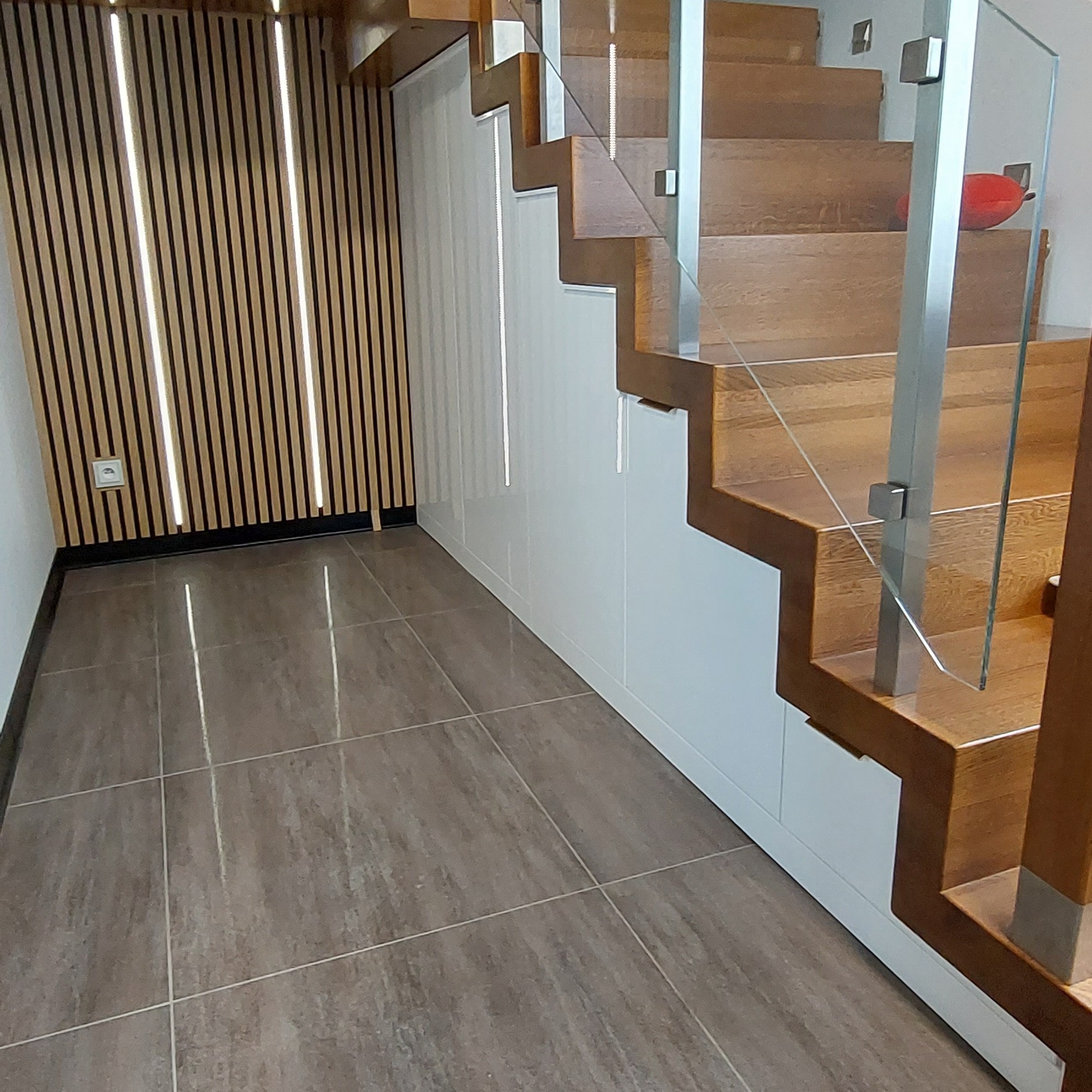 Nowoczesne drewniane schody z szklaną balustradą, zintegrowane z białą zabudową meblową, na podłodze szare płytki, dekoracyjna ściana z lameli z oświetleniem.