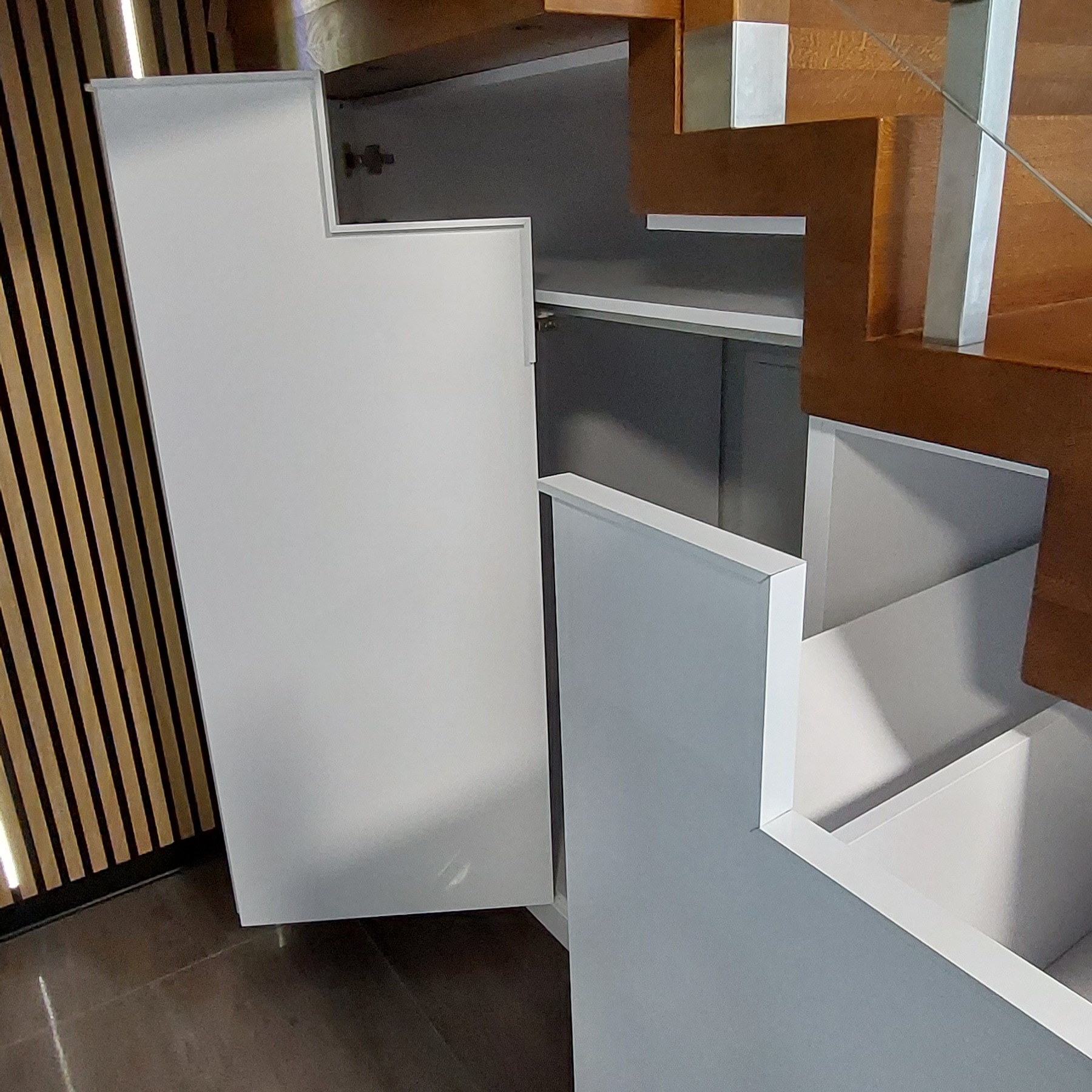 Biała szafa wnękowa pod schodami z otwartymi drzwiczkami, widoczne półki wewnątrz, obok drewniane schody z metalowymi elementami.