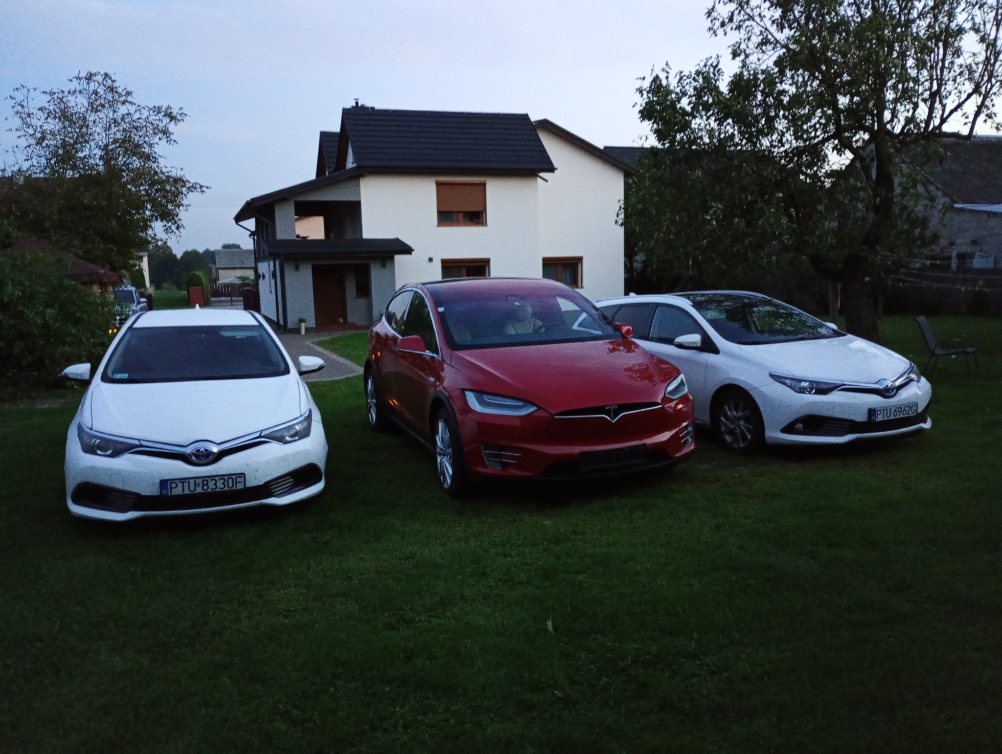 Trzy samochody osobowe (białe Toyoty i czerwona Tesla) zaparkowane na trawniku przed domem jednorodzinnym o zmierzchu.