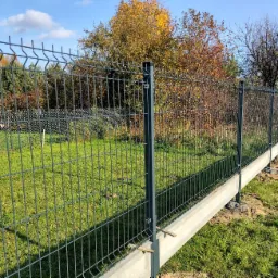 Nowoczesne, szare ogrodzenie panelowe z podmurówką betonową na trawiastym terenie, widok z boku, słoneczny dzień.