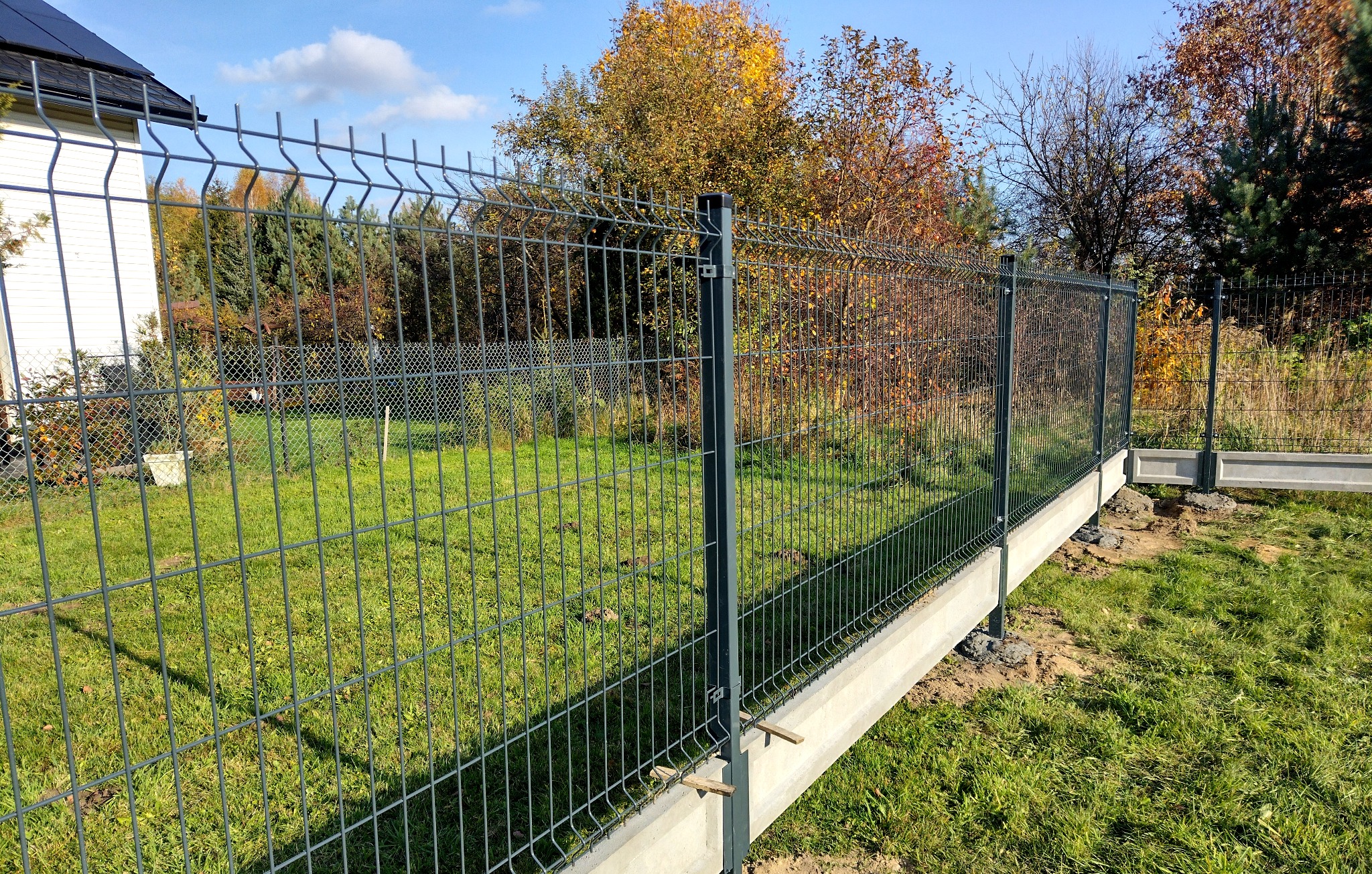 Nowoczesne, szare ogrodzenie panelowe z podmurówką betonową na trawiastym terenie, widok z boku, słoneczny dzień.