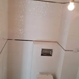 Nowoczesna łazienka z białymi, trójwymiarowymi płytkami na ścianach, szarą podłogą i podwieszaną toaletą. Widoczny fragment instalacji elektrycznej na suficie.