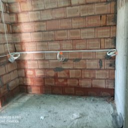 Szymon Kafka - Surowy stan wnętrza z widocznymi przewodami elektrycznymi zamocowanymi do ściany z czerwonej cegły. Na przewodach pomarańczowe znaczniki.