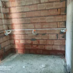 Surowy stan wnętrza z widocznymi przewodami elektrycznymi zamocowanymi do ściany z czerwonej cegły. Na przewodach pomarańczowe znaczniki.