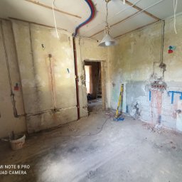 Szymon Kafka - Wnętrze w trakcie remontu: widoczne odsłonięte ściany z instalacjami elektrycznymi i hydraulicznymi, poziomica oparta o ścianę, surowe wykończenie, lampa wisząca, widoczne przewody i kable.