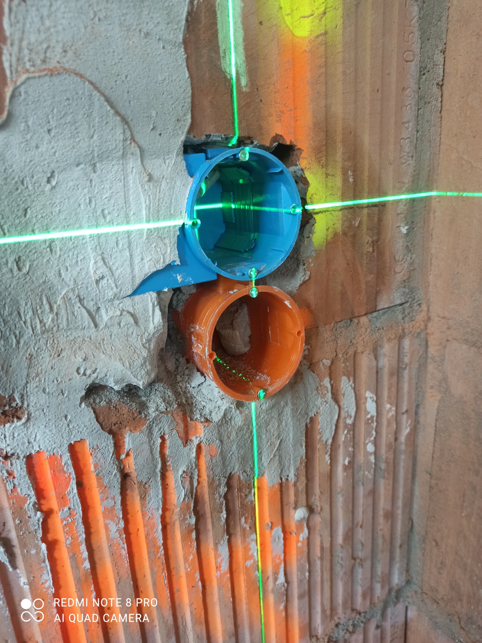 Niebieska i pomarańczowa puszka instalacyjna zamontowana w surowej ścianie z cegły, wyznaczona laserowo w pionie i poziomie.