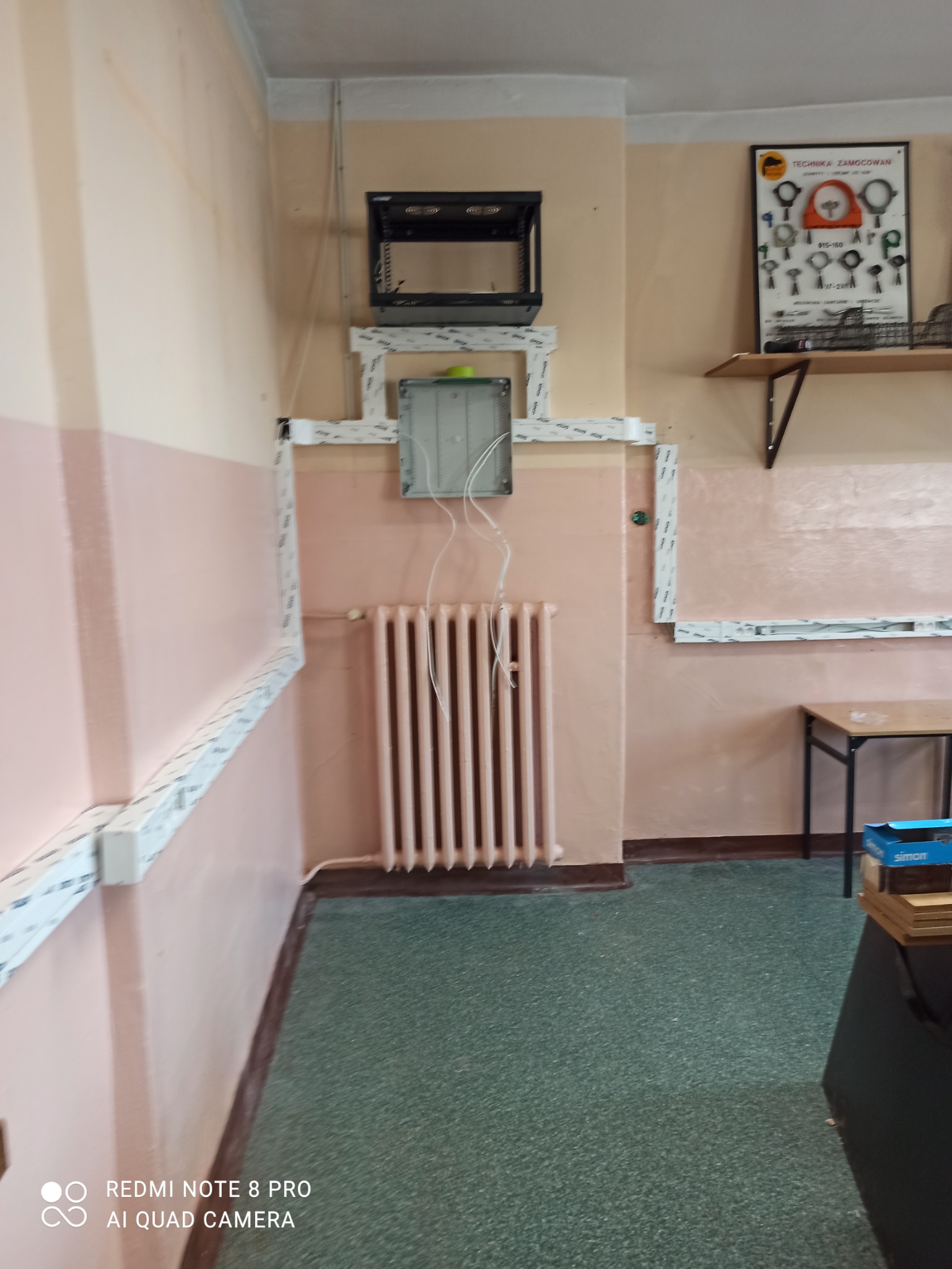 Szare korytka kablowe na ścianie w budynku, z puszką podłączeniową i wiszącymi przewodami nad grzejnikiem, oraz zamontowana szafa rack powyżej.