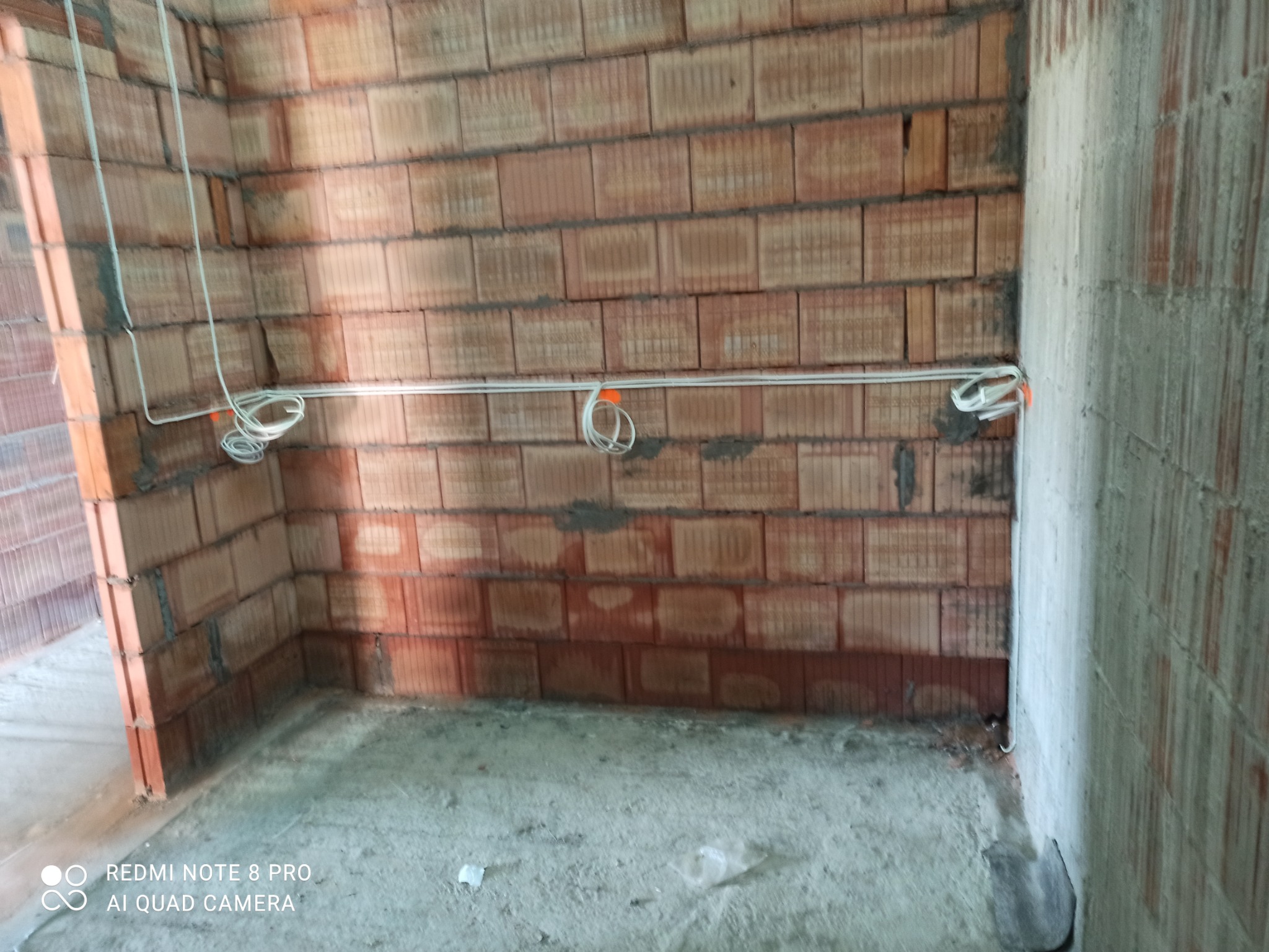 Surowy stan wnętrza z widocznymi przewodami elektrycznymi zamocowanymi do ściany z czerwonej cegły. Na przewodach pomarańczowe znaczniki.