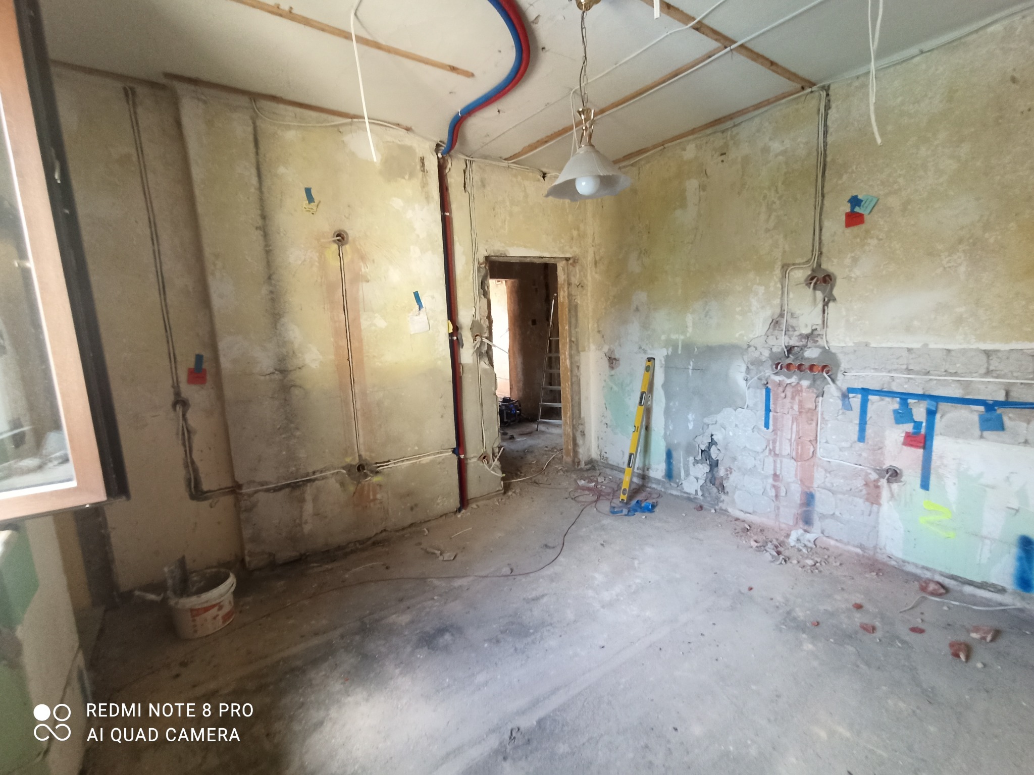 Wnętrze w trakcie remontu: widoczne odsłonięte ściany z instalacjami elektrycznymi i hydraulicznymi, poziomica oparta o ścianę, surowe wykończenie, lampa wisząca, widoczne przewody i kable.