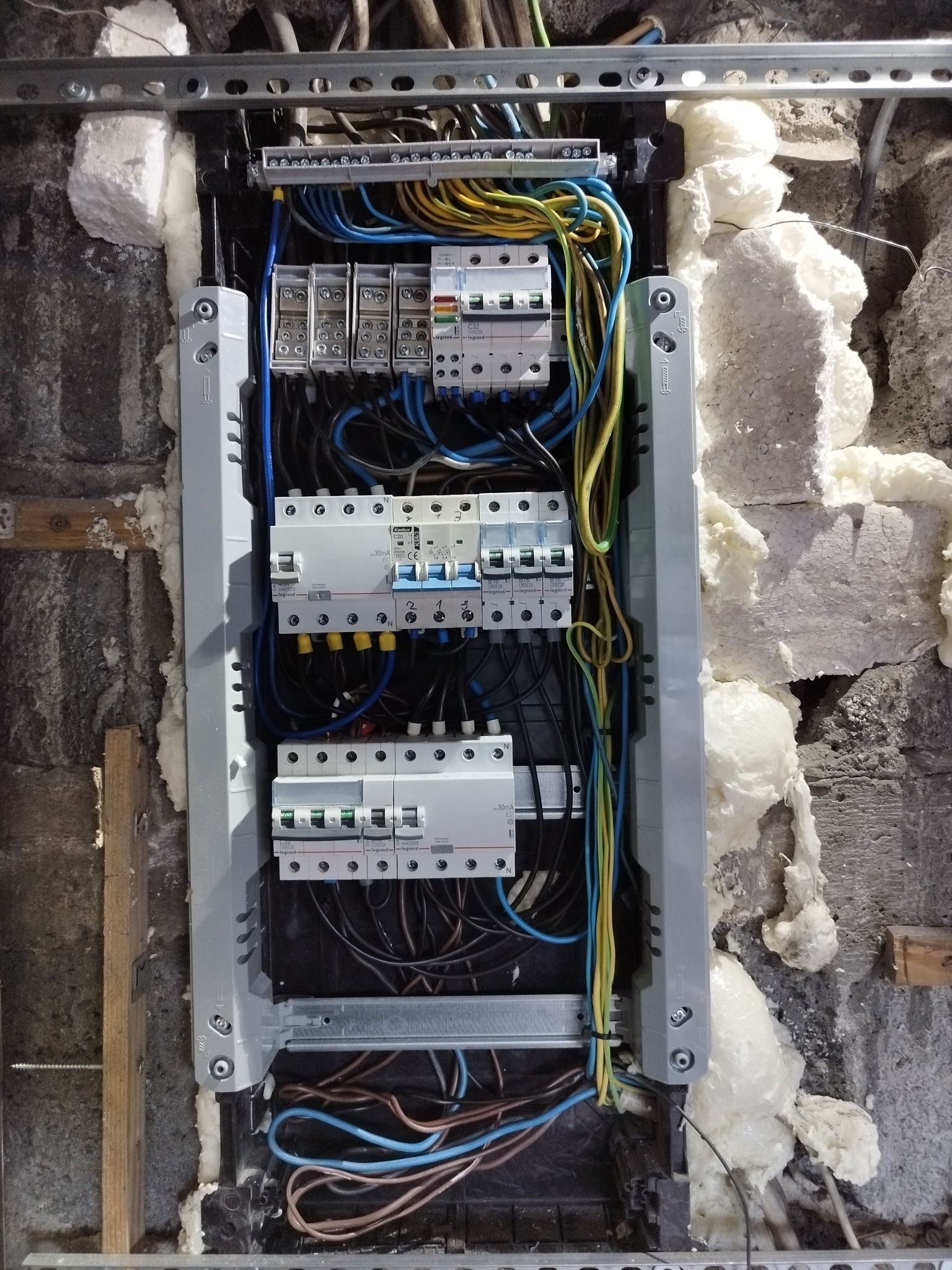 Rozdzielnica elektryczna z zainstalowanymi bezpiecznikami Legrand i Kanlux, widoczne przewody w kolorach niebieskim, żółto-zielonym i czarnym, zamontowana w ścianie z izolacją piankową.