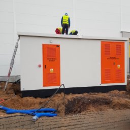 EL-MAR Marek Cegielski - Montaż komory transformatora i rozdzielni nN 0,4kV przez dwóch elektryków w odblaskowych kamizelkach na dachu kontenerowej stacji transformatorowej, widoczne kable w ziemi i drabina oparta o budynek.