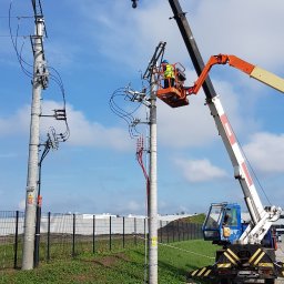 EL-MAR Marek Cegielski - Pracownik w podnośniku koszowym, w żółtej kamizelce i niebieskim kasku, montuje kable na słupie energetycznym. Obok widoczny drugi słup z już podłączonymi kablami. W tle błękitne niebo z chmurami...