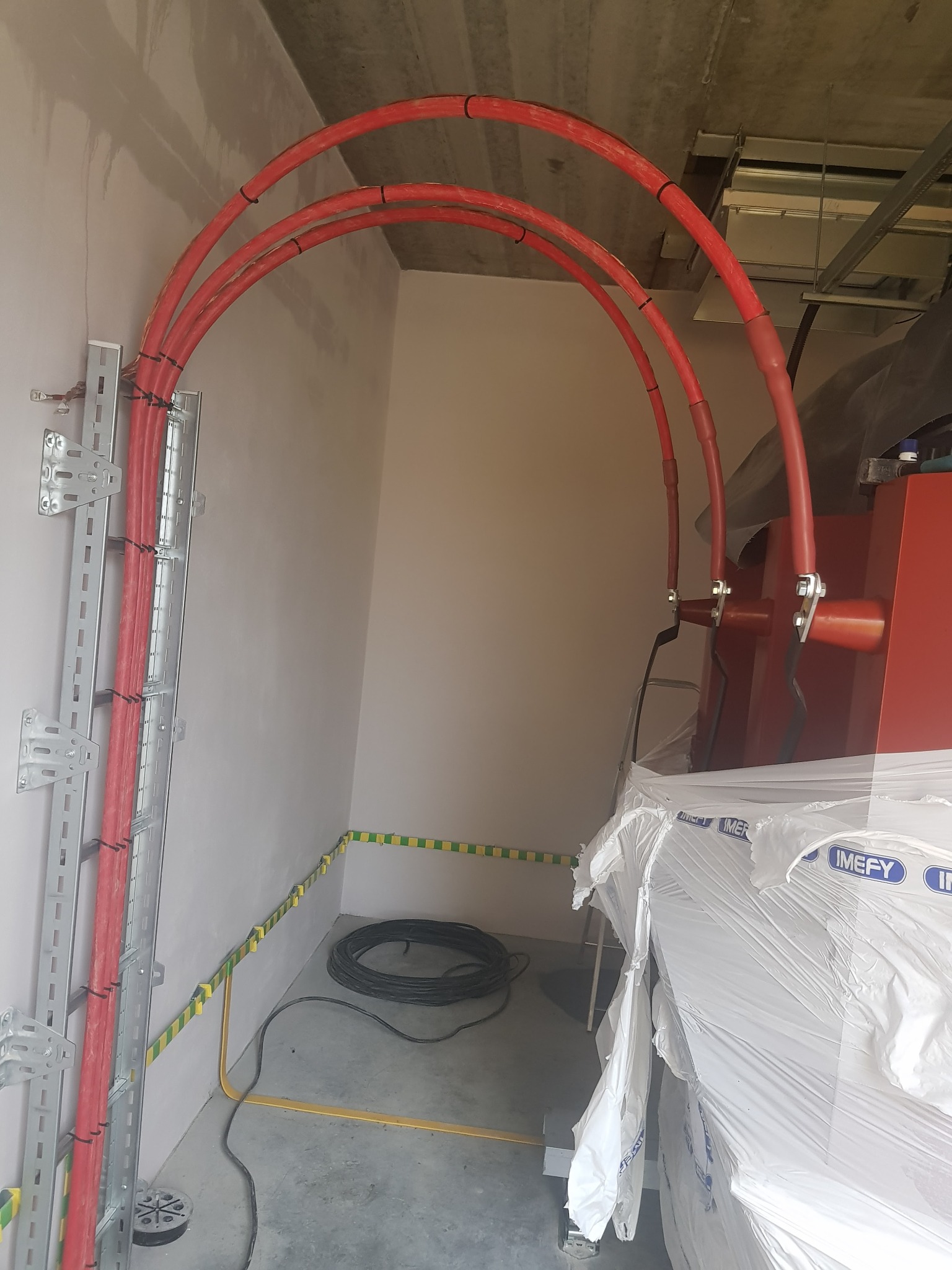 Czerwone kable elektryczne prowadzone po metalowej drabince przy ścianie, podłączone do urządzenia w tle, z widocznym uziemieniem i zwitkiem kabla na podłodze.