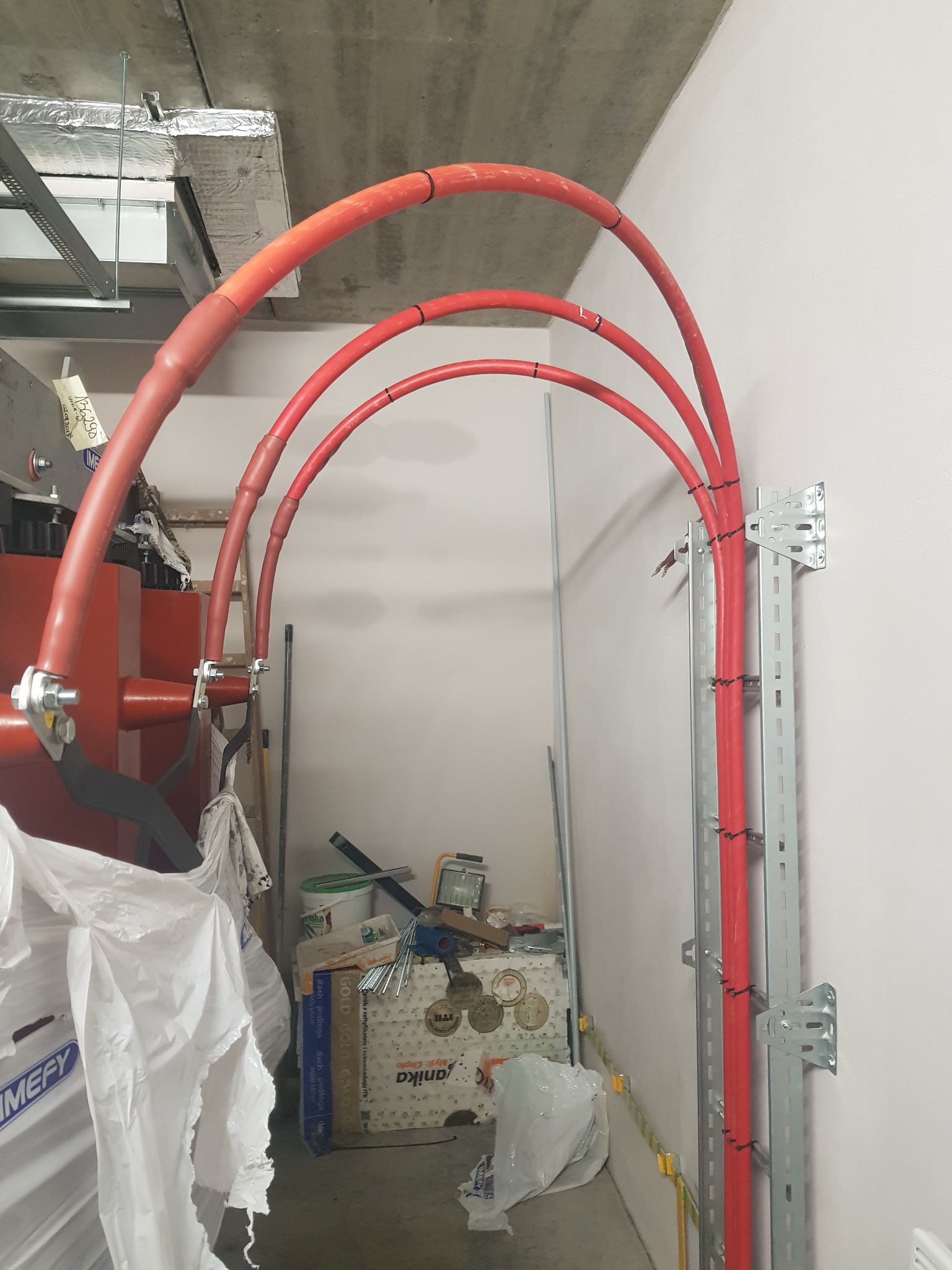 Trzy czerwone, zaizolowane kable prowadzone łukiem od ściany do urządzenia elektrycznego, zamocowane na metalowych wspornikach, widoczne narzędzia i materiały budowlane w tle.