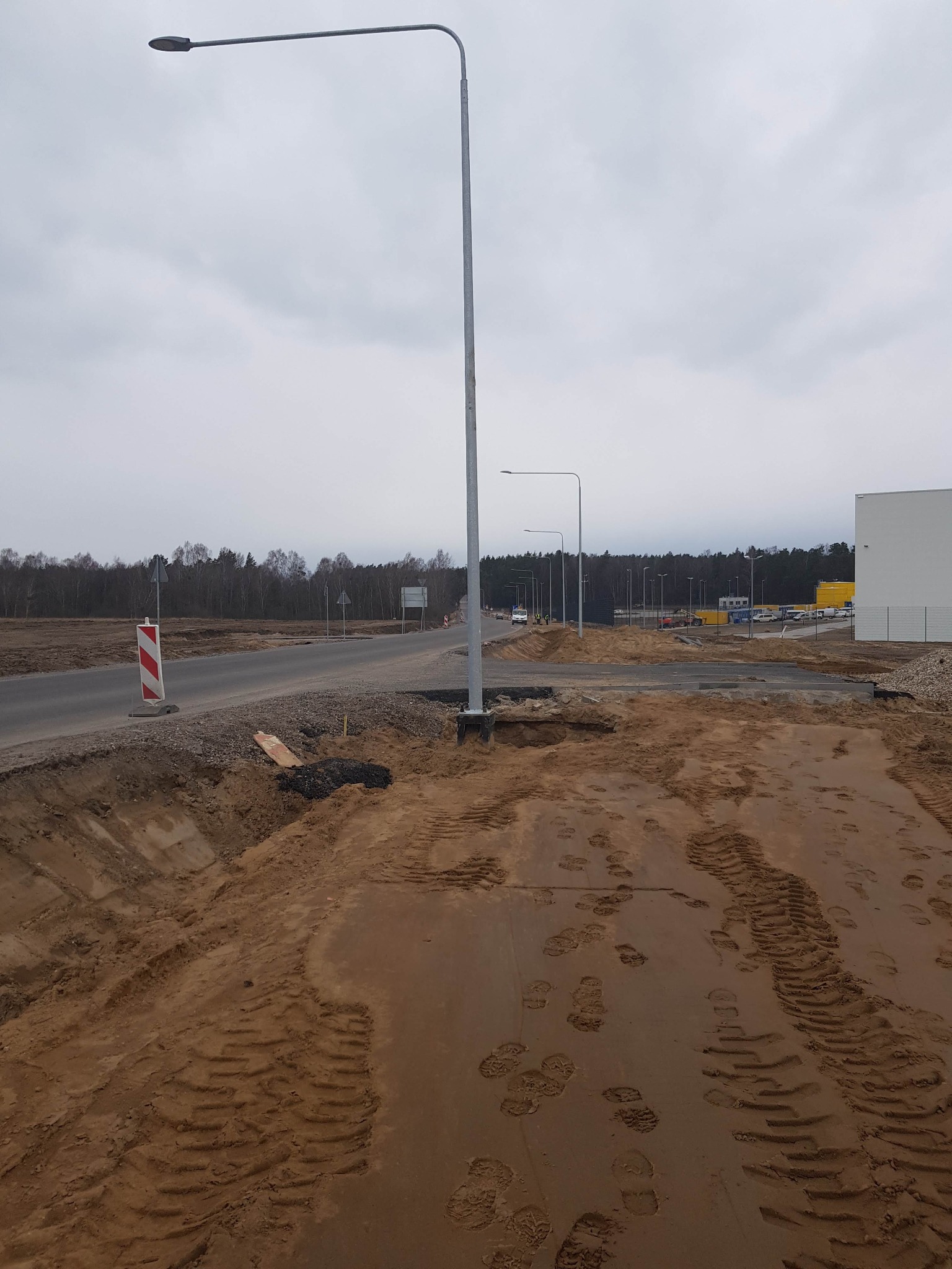 Montaż nowych, szarych latarni ulicznych na placu budowy z widocznymi śladami opon i butów w ziemi.