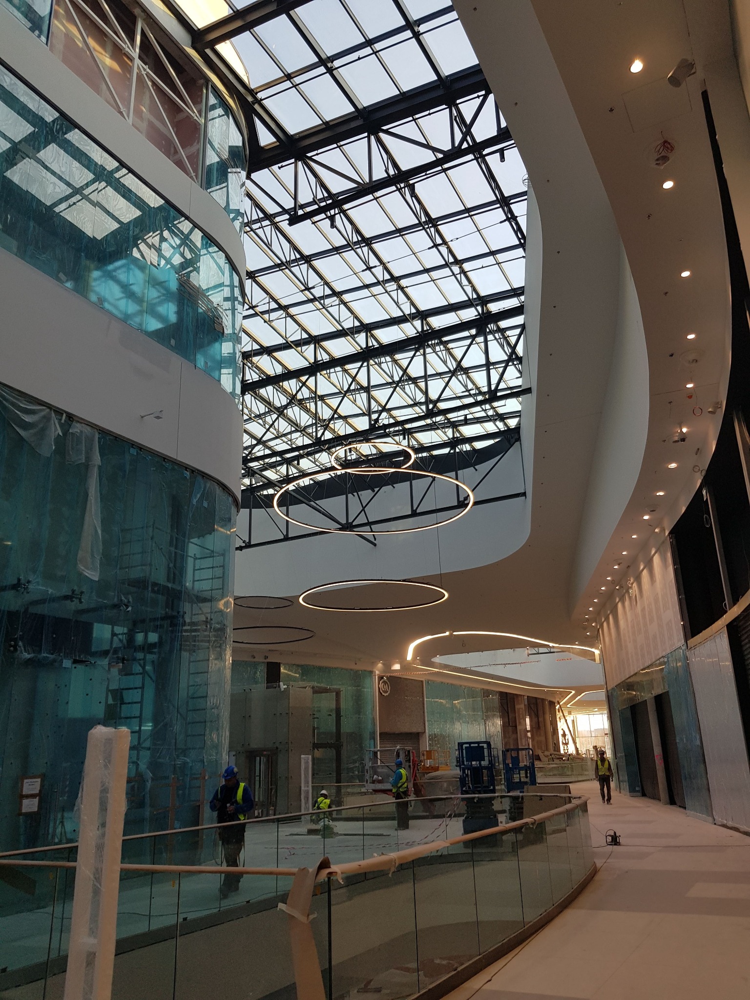 Wnętrze nowoczesnego centrum handlowego w trakcie budowy z widocznymi okrągłymi lampami pod szklanym dachem i pracownikami w kaskach.