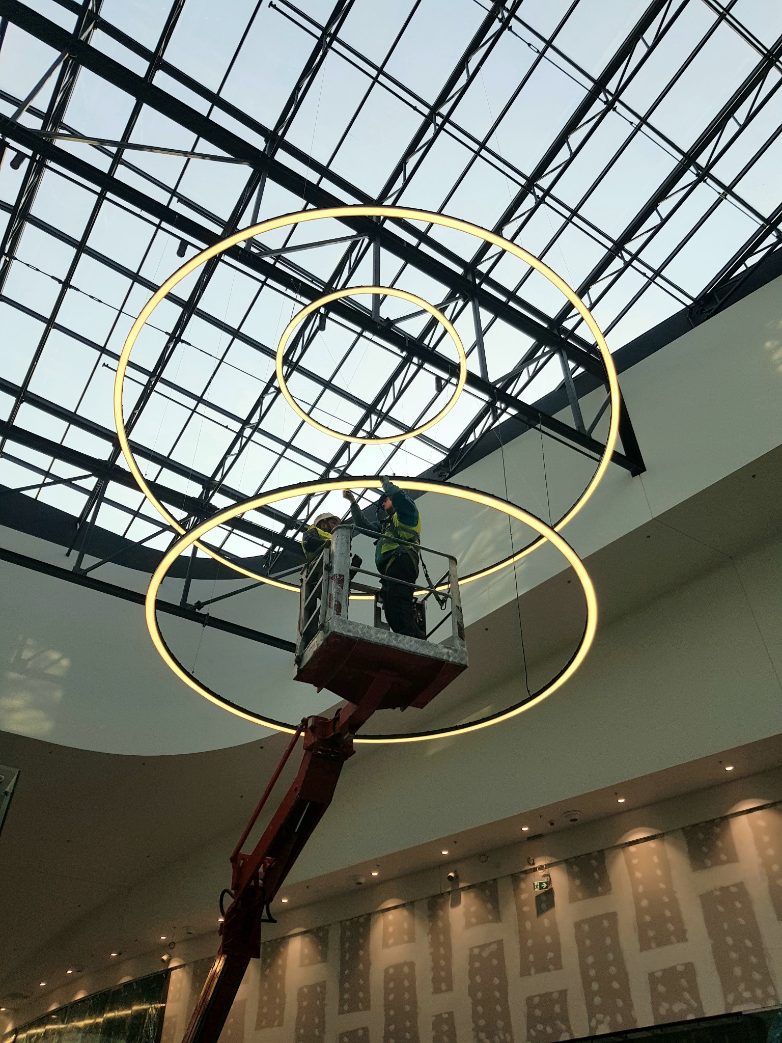 Montaż potrójnego, okrągłego oświetlenia LED w galerii handlowej z przeszklonym dachem; dwóch pracowników na podnośniku koszowym w żółtych kamizelkach i kaskach.