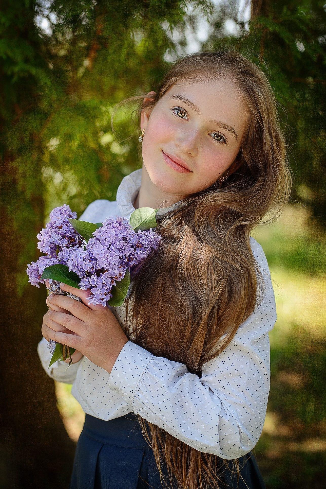 Portret uśmiechniętej, młodej dziewczyny trzymającej bukiet bzu w dłoniach, na tle zieleni drzew.