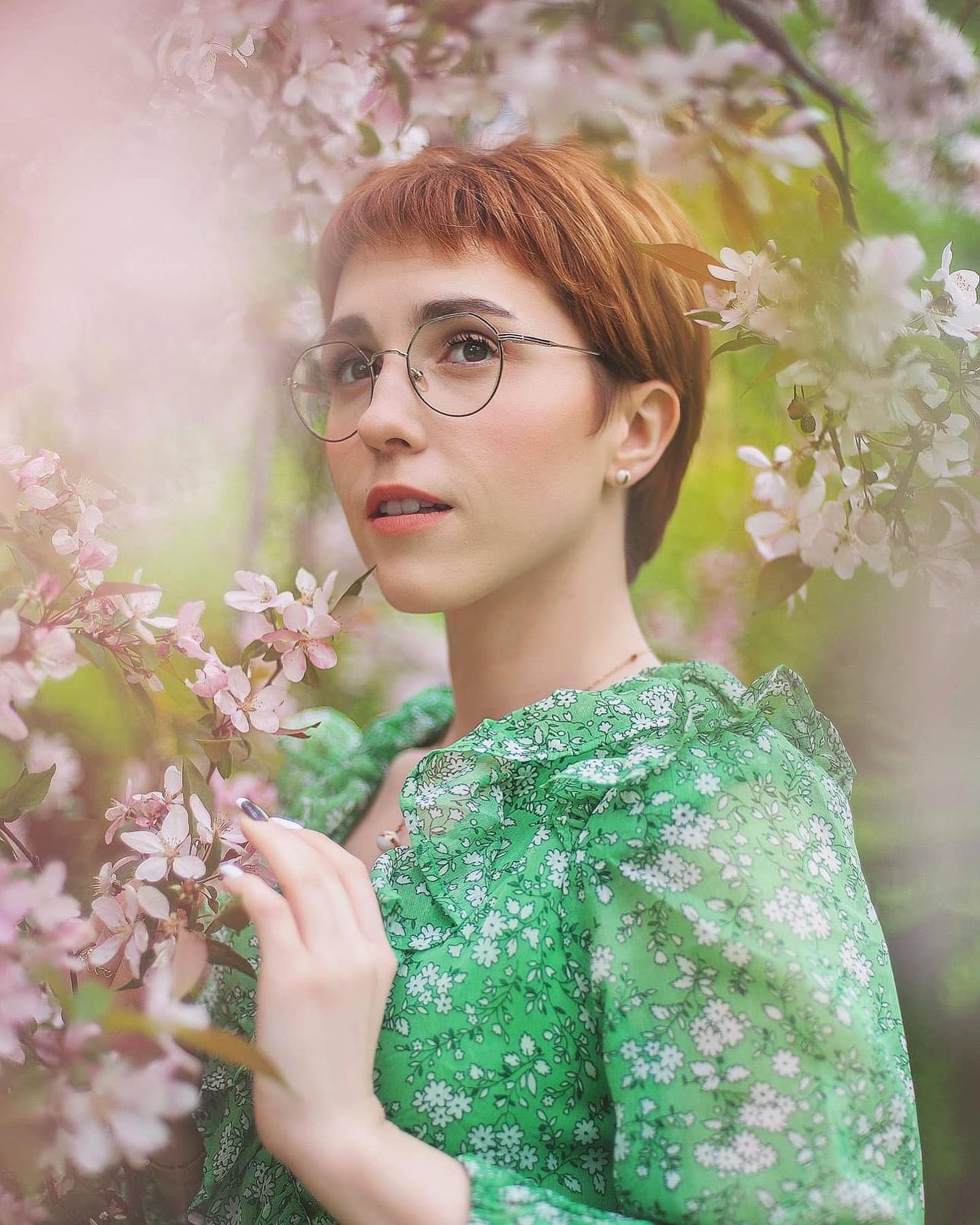 Portret kobiety w okularach, z krótkimi rudymi włosami, ubrana w zieloną sukienkę w białe kwiaty, pozująca wśród kwitnących gałęzi drzewa owocowego, z delikatnym uśmiechem i pomalowanymi paznokciami.