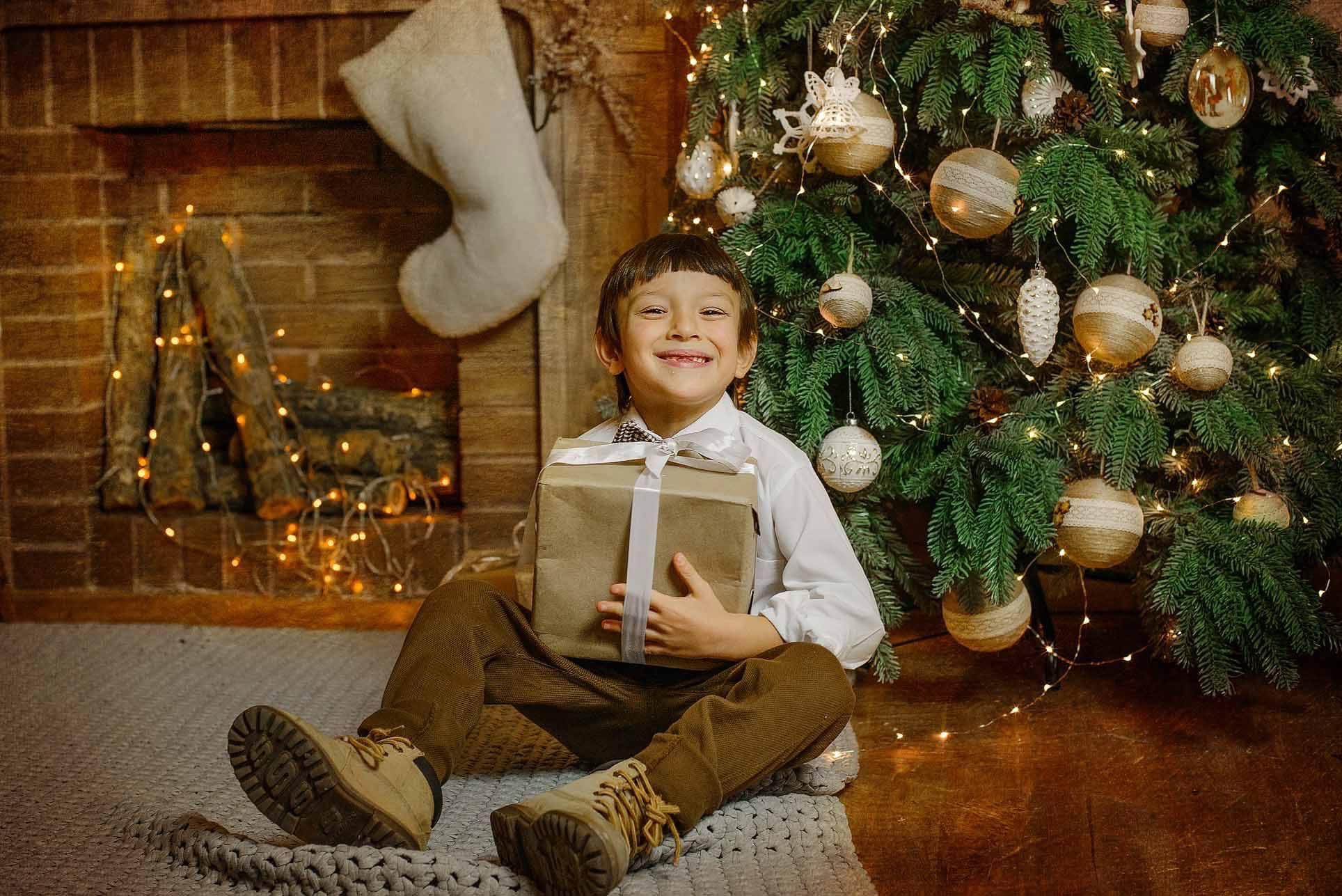 Uśmiechnięty chłopiec w brązowych spodniach i białej koszuli siedzi pod choinką, trzymając prezent owinięty brązowym papierem i przewiązany białą wstążką, w tle kominek z dekoracjami świątecznymi.