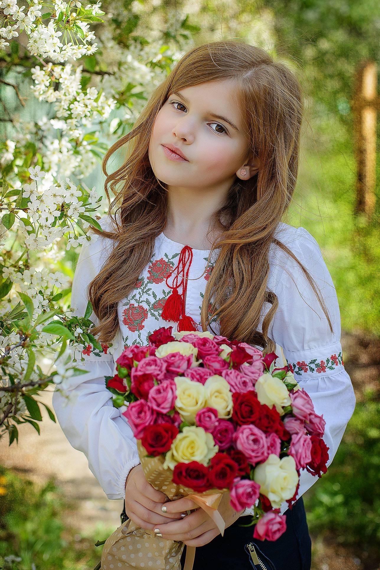Dziewczynka w ludowej bluzce z haftem trzyma bukiet czerwonych, różowych i żółtych róż na tle kwitnącej wiśni.