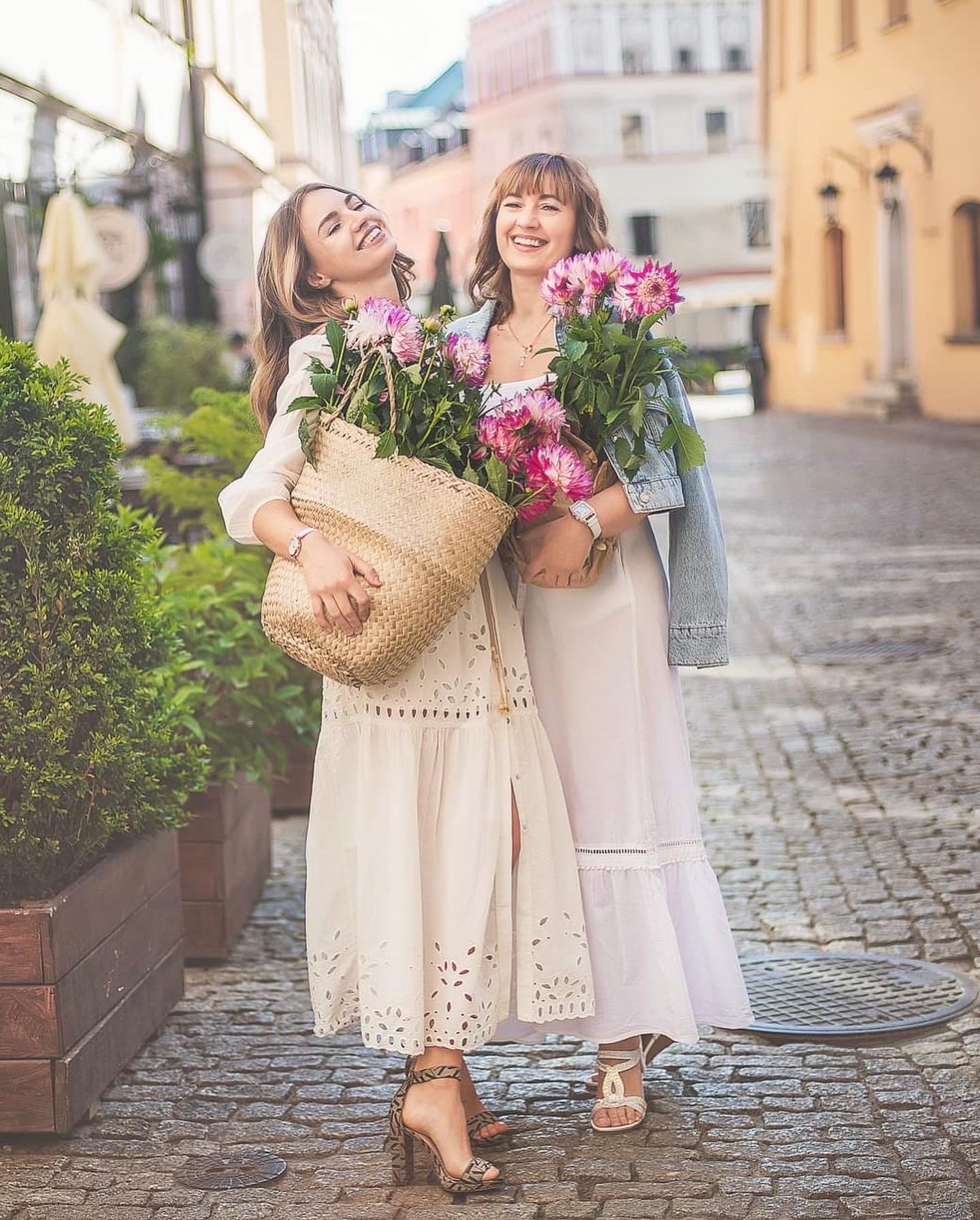 Dwie uśmiechnięte kobiety w zwiewnych białych sukienkach, trzymające bukiety różowych dalii i wiklinowy koszyk, pozują na brukowanej uliczce w słoneczny dzień.