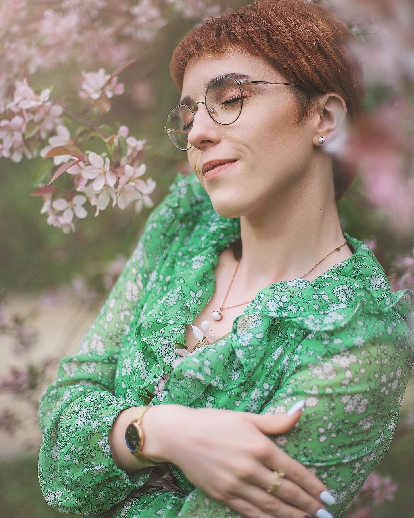 Portret kobiety w okularach, w zielonej sukience w kwiaty, stojącej na tle kwitnącego drzewa, z delikatnym uśmiechem i zamkniętymi oczami, z widoczną biżuterią: kolczyki, naszyjnik i zegarek.