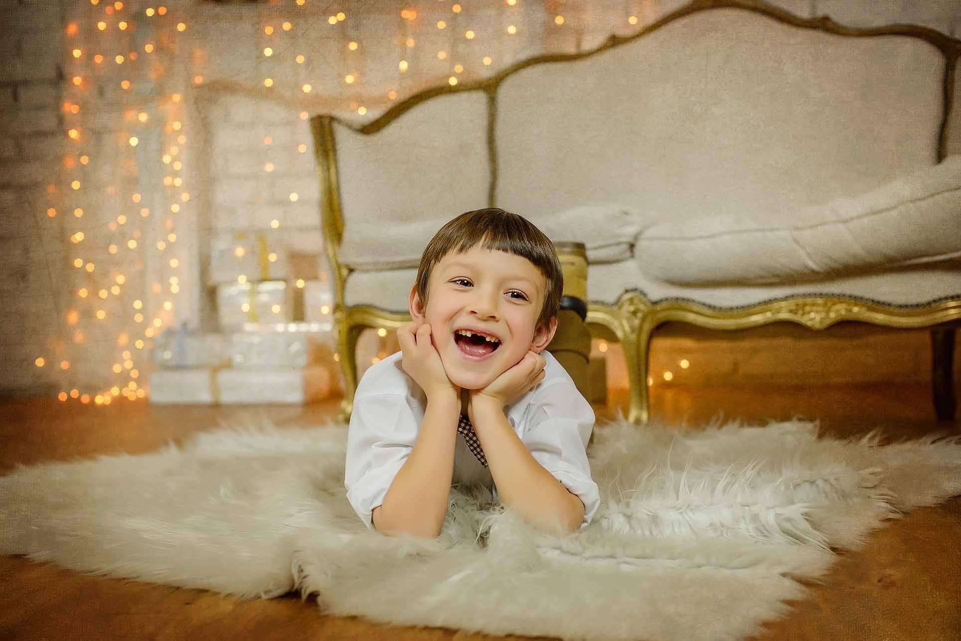Uśmiechnięty chłopiec leży na puszystym, białym dywanie, opierając brodę na dłoniach, w tle kremowa sofa z pozłacanymi zdobieniami, prezenty i girlanda światełek.