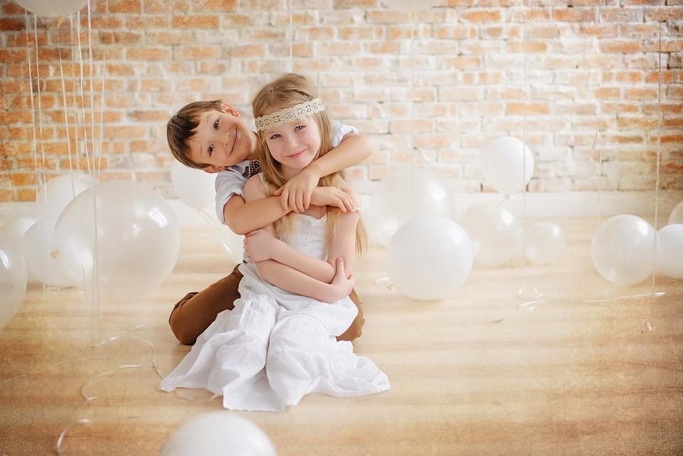 Uśmiechnięty chłopiec obejmuje siedzącą dziewczynkę w białej sukience i z opaską na głowie, otoczeni białymi balonami na tle ceglanej ściany.