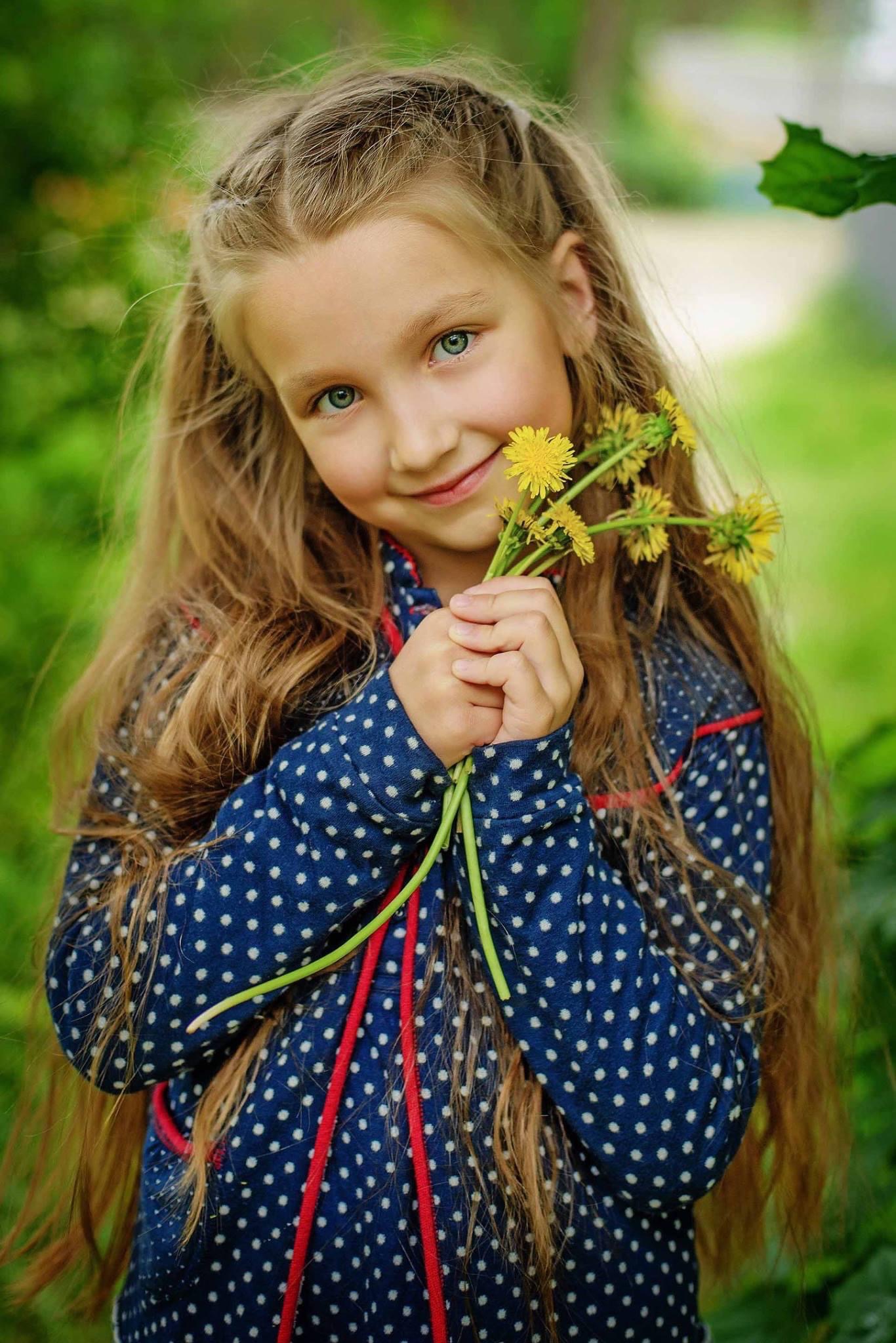 Uśmiechnięta dziewczynka z długimi, blond włosami i niebieskimi oczami trzyma bukiecik żółtych mleczy na tle zieleni.