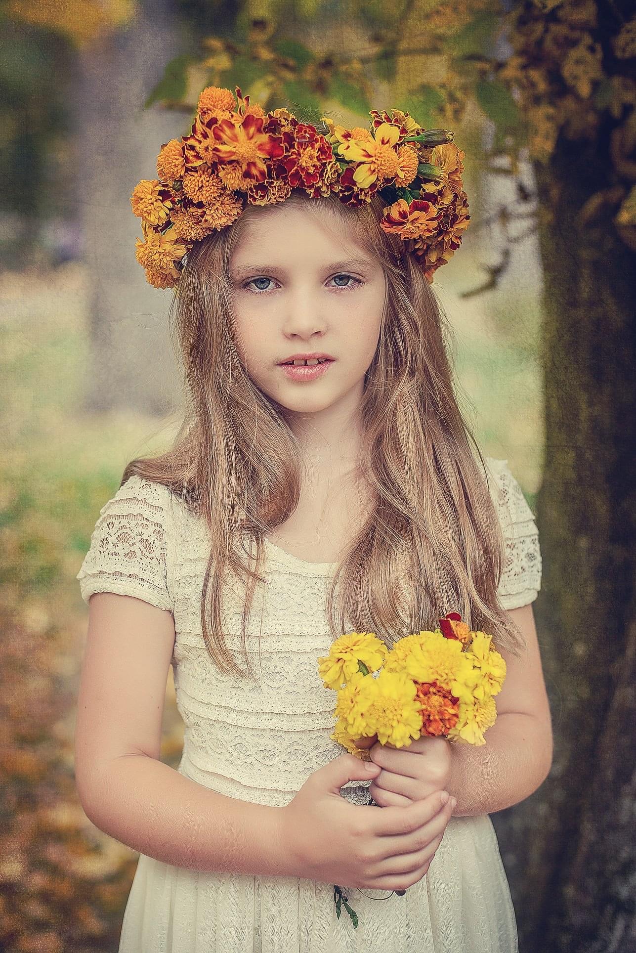 Portret dziewczynki z wiankiem z nagietków na głowie, trzymającej bukiecik żółtych kwiatów na tle jesiennego parku.