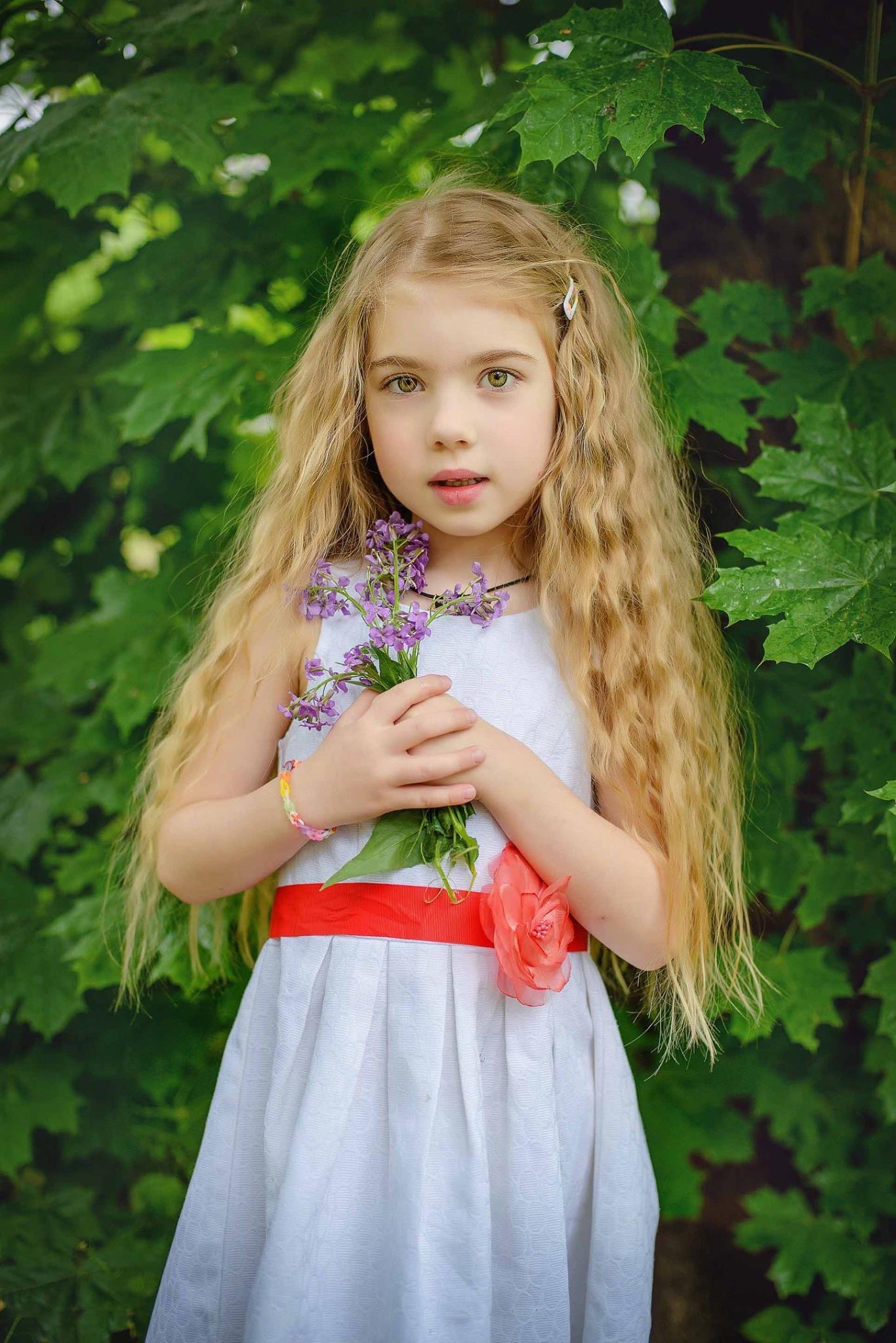 Portret dziewczynki z długimi blond włosami trzymającej bukiet fioletowych kwiatów na tle zielonych liści, ubrana w białą sukienkę z czerwonym paskiem i ozdobnym kwiatem.