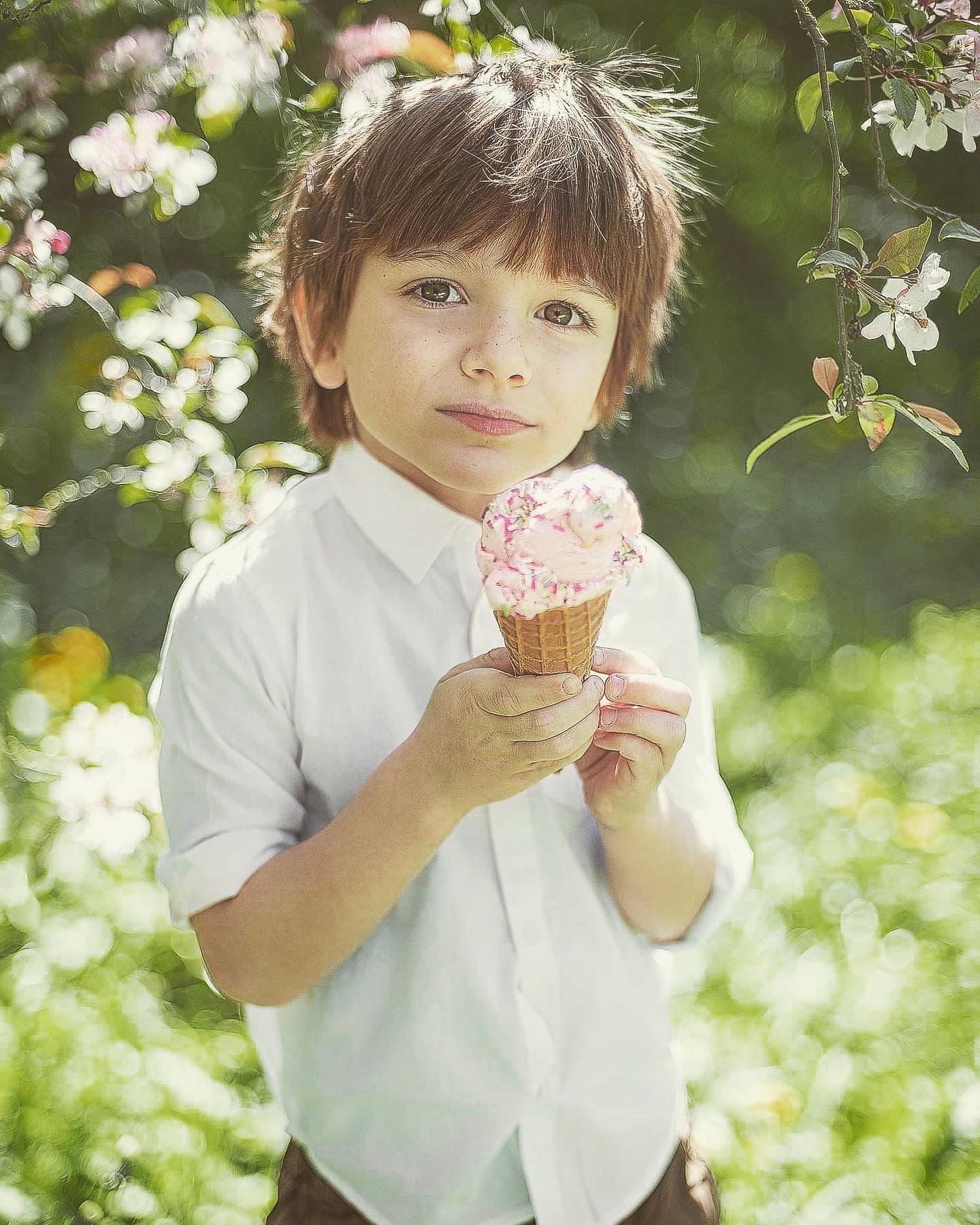 Chłopiec z piegami, w białej koszuli, trzyma w dłoniach różowy lód w wafelku na tle kwitnących gałęzi drzewa.