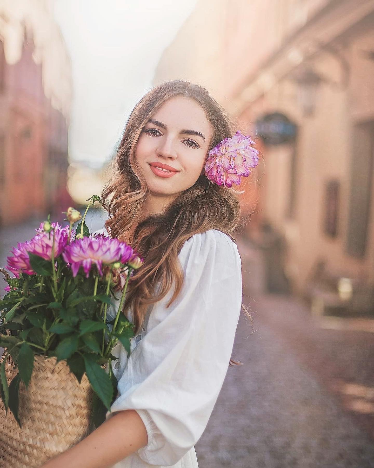 Portret uśmiechniętej młodej kobiety z kwiatem we włosach i bukietem dalii w wiklinowym koszu, na tle uliczki starego miasta.