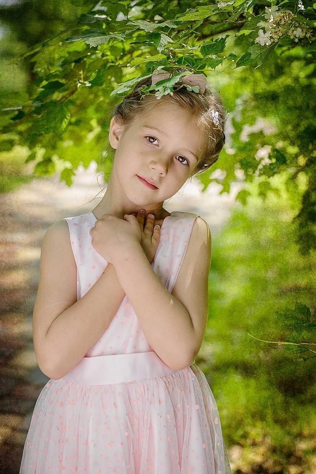 Portret uroczej dziewczynki w różowej sukience, z dłońmi złożonymi pod szyją, pozującej pod zielonymi gałęziami drzewa w naturalnym świetle.