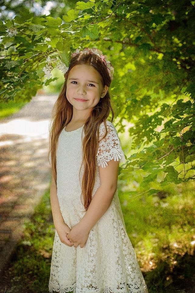 Portret uśmiechniętej dziewczynki w koronkowej sukience i wianku, stojącej pod drzewem w słoneczny dzień.