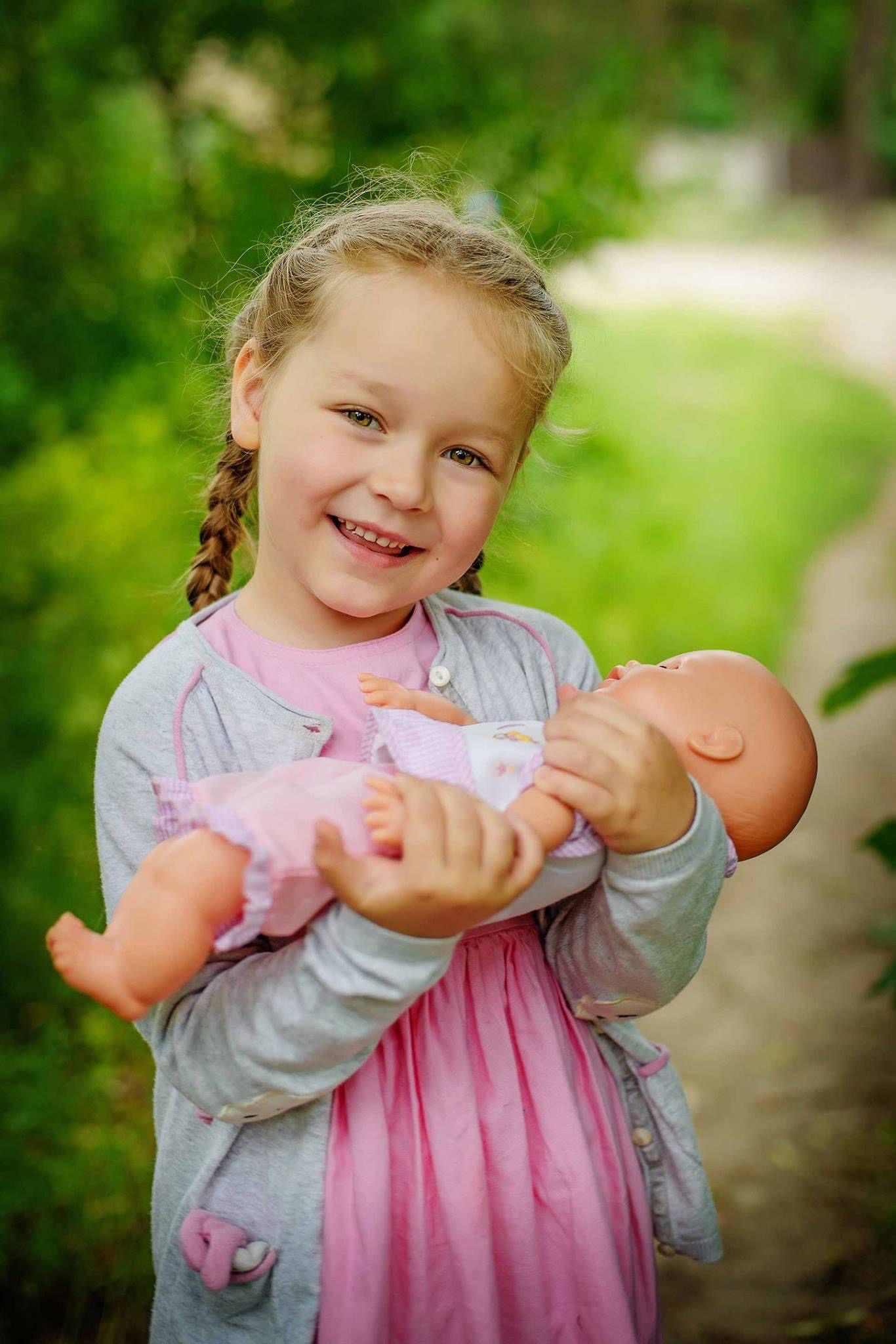 Uśmiechnięta dziewczynka z warkoczami trzyma lalkę w różowej sukience na tle zieleni.