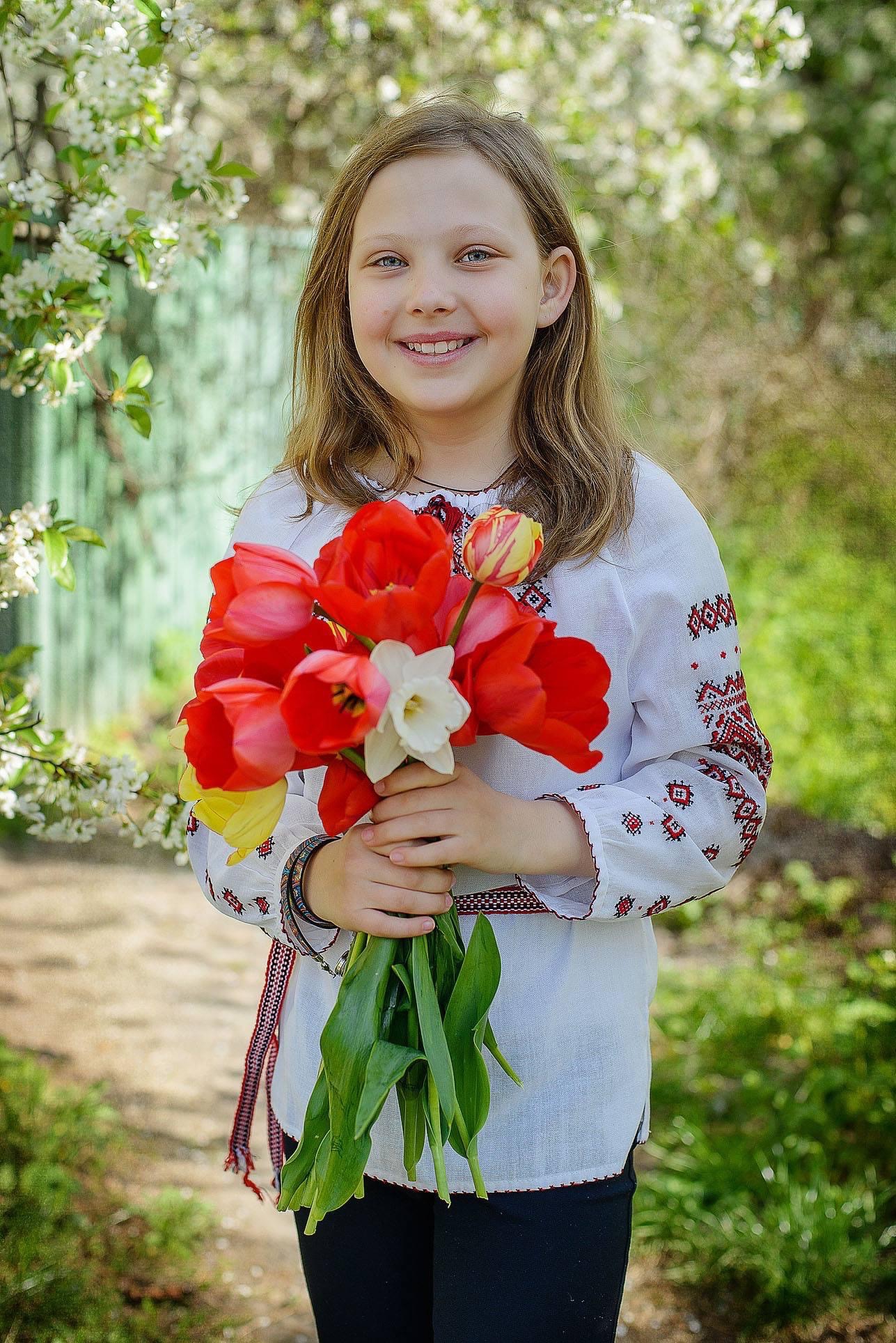 Uśmiechnięta dziewczynka w haftowanej bluzce trzyma bukiet czerwonych tulipanów i białego narcyza na tle kwitnących drzew